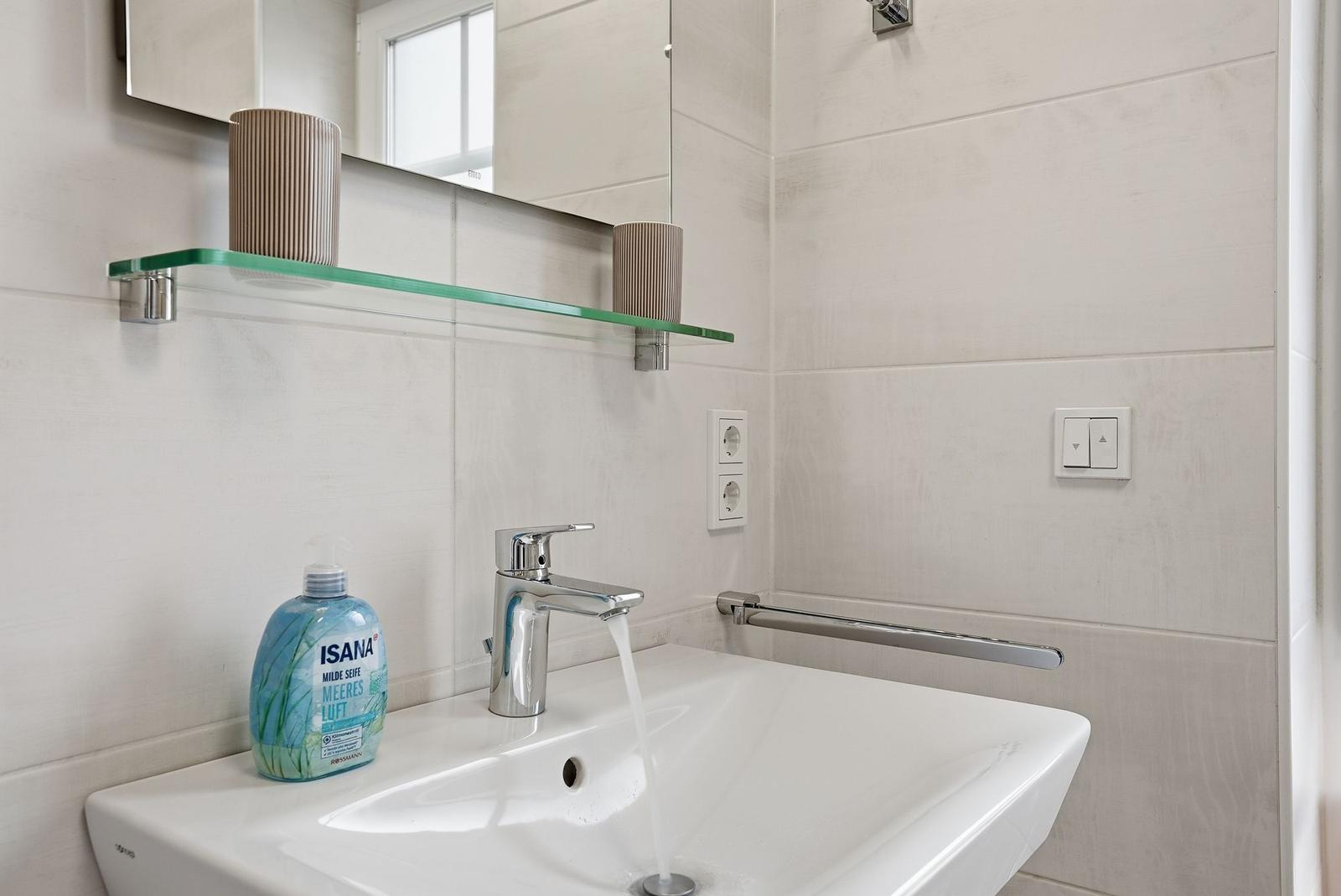 Waschbecken mit laufendem Wasser, Spiegel und Glasregal mit Behältern.