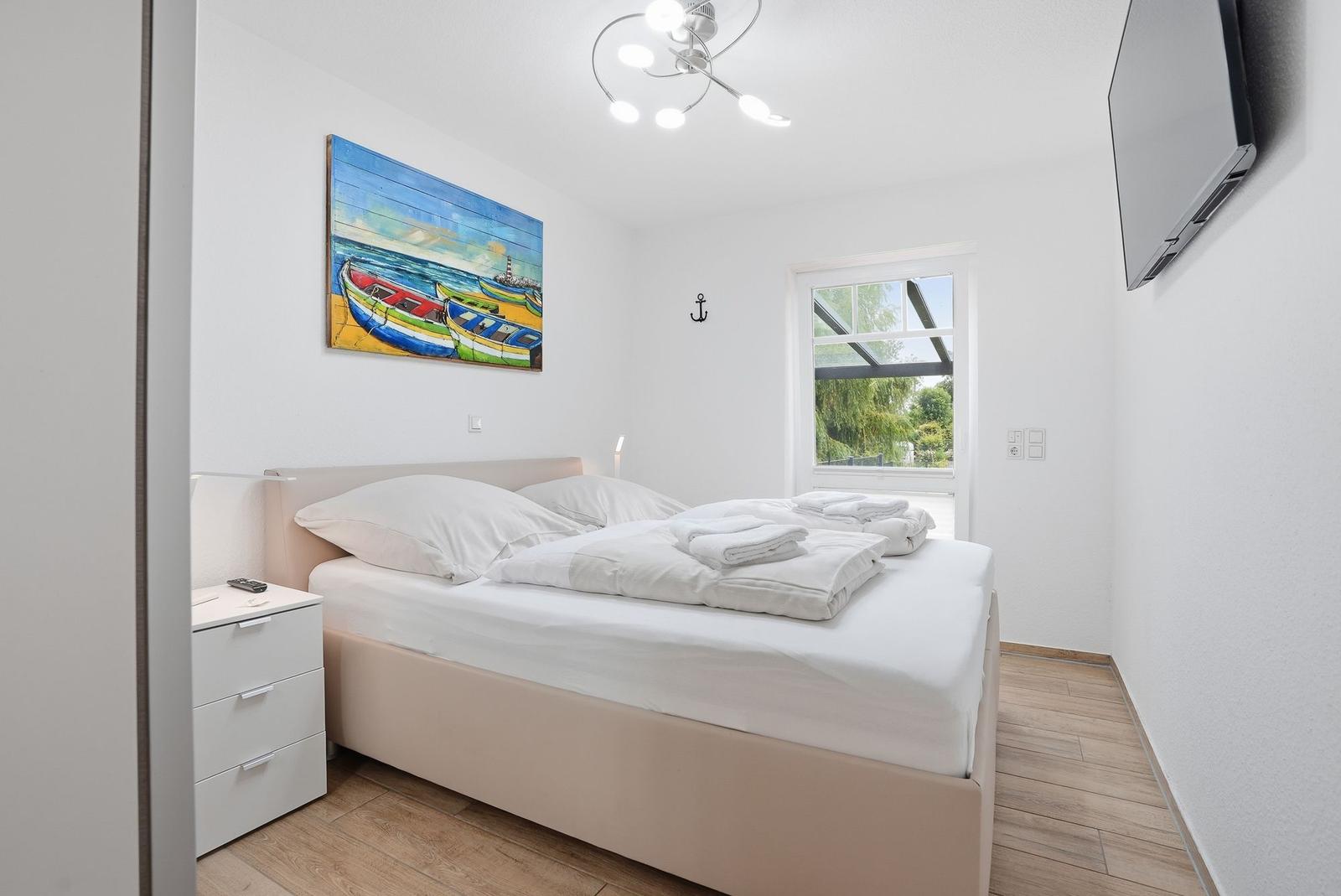Schlafzimmer mit Bett, Nachttisch und Wandbild. Fenster zeigt Garten. TV an der Wand.