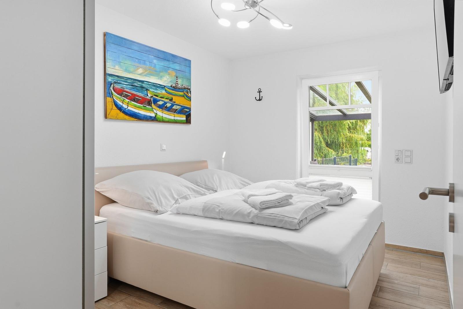 Schlafzimmer mit Bett, Wandbild und Fenster zum Garten.