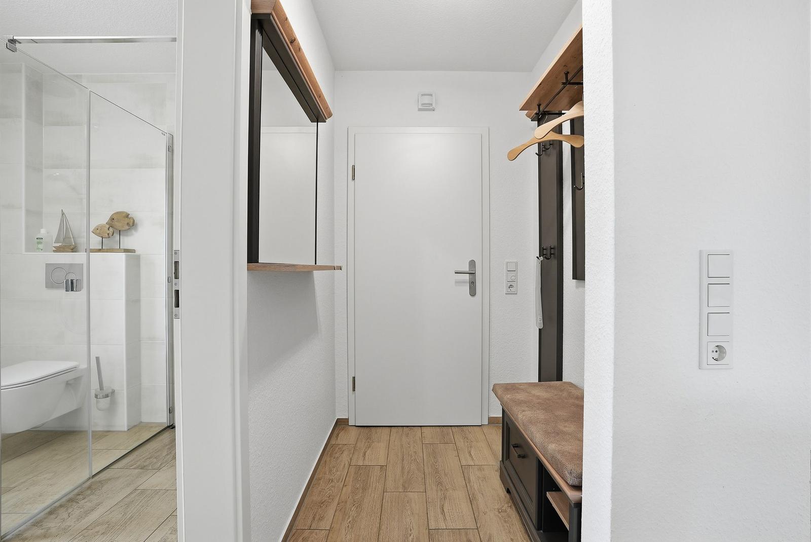 Flur mit Holzboden, Kleiderhaken und Sitzbank. Bad mit Dusche und Toilette sichtbar.