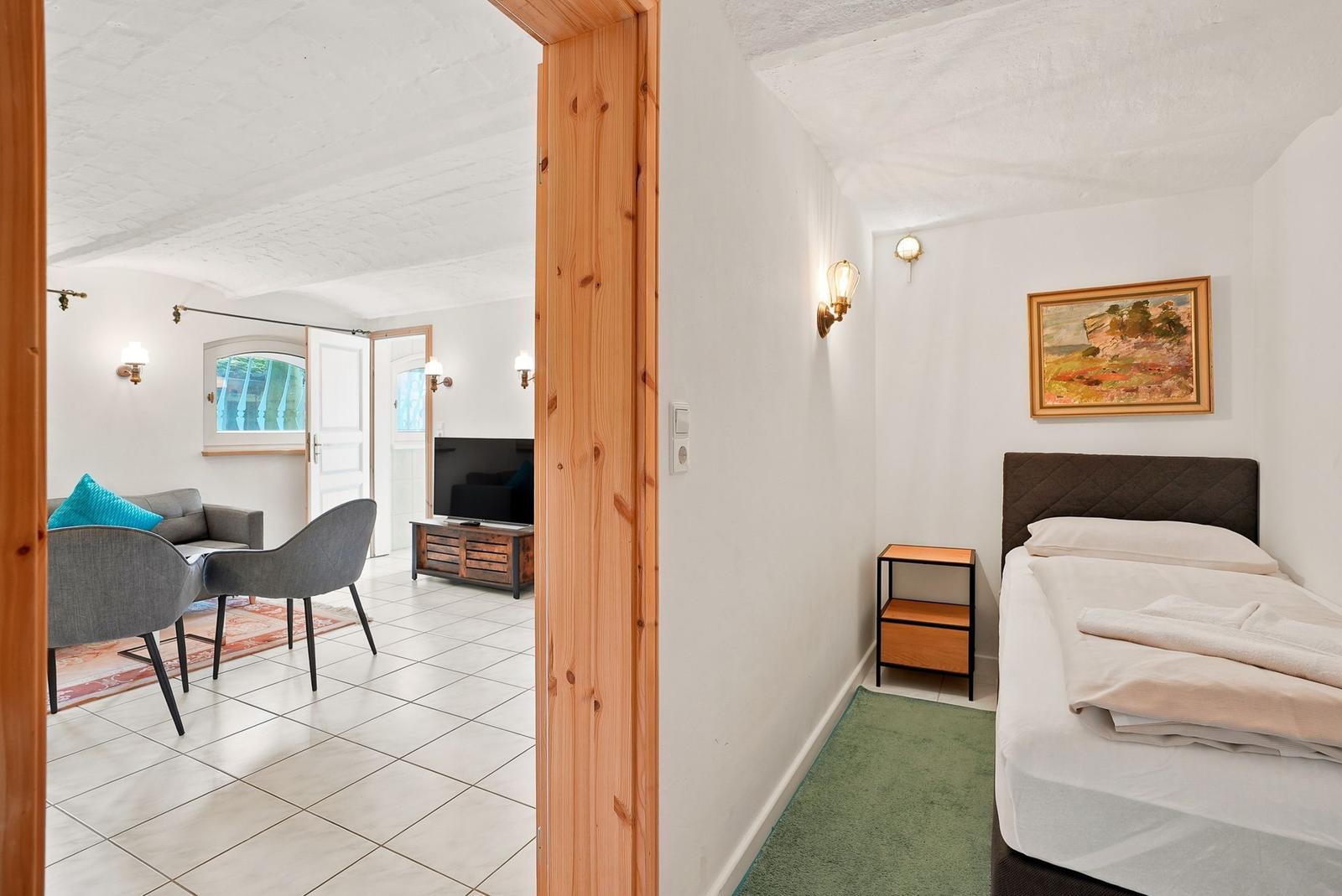 Schlafzimmer mit Bett und Nachttisch. Durchgang zum Wohnzimmer mit Sofa und Fernseher.