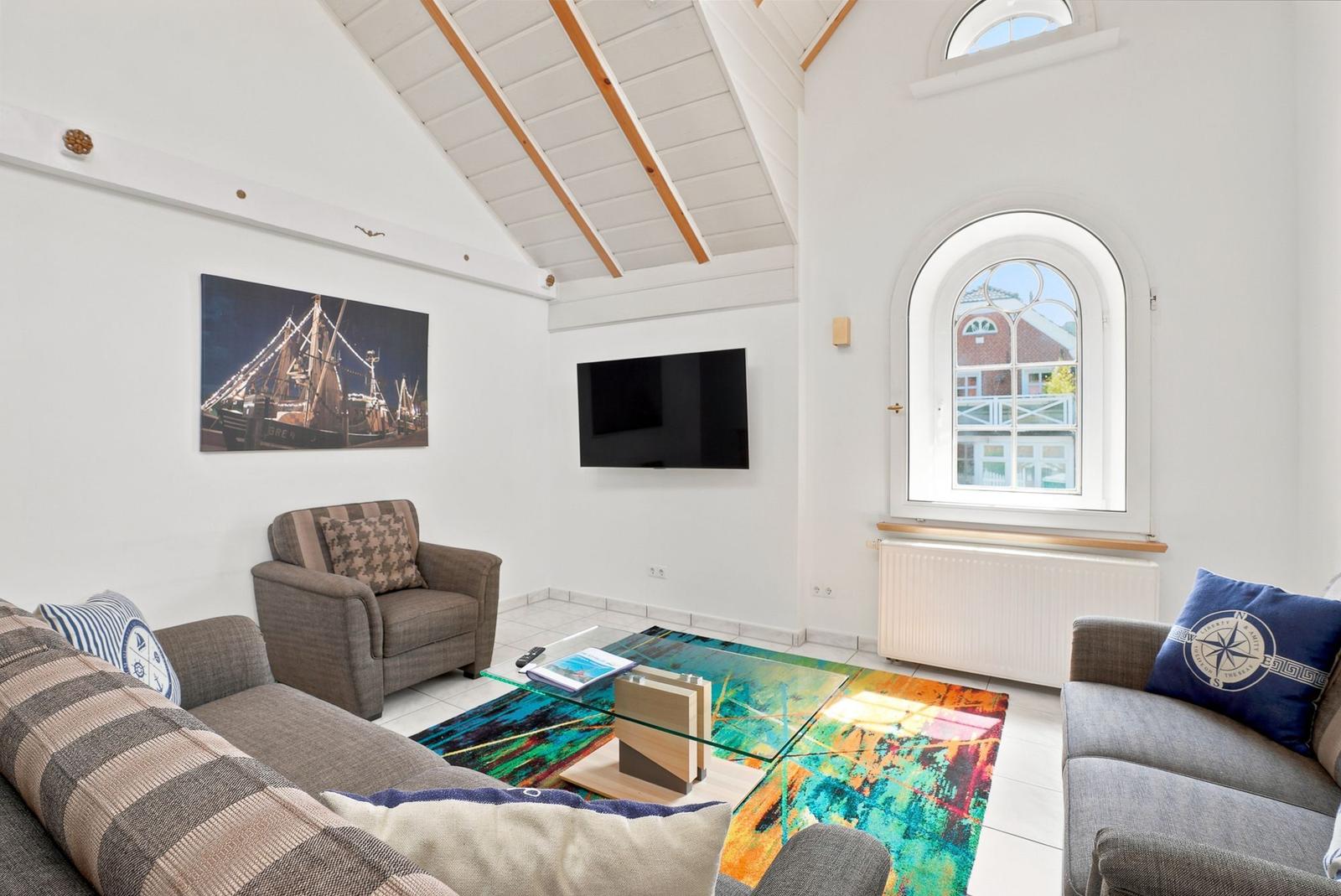 Wohnzimmer mit Sofa, Sessel, Glas-Tisch und Wandbild. Hochdecke mit Holzbalken und Bogenfenster.
