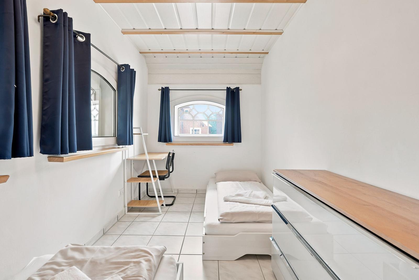 Zimmer mit Bett, Schreibtisch, Kleiderschrank und Fenster mit blauen Vorhängen.