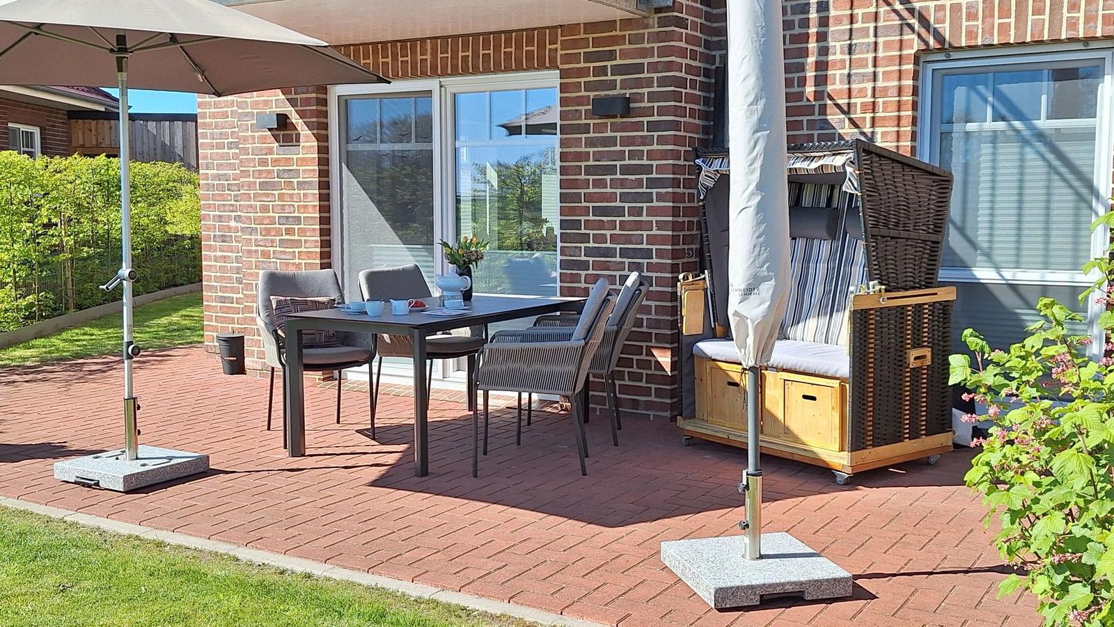 Terrasse mit Tisch, Stühlen und Sonnenschirmen. Gartenhaus und Blumen sind sichtbar.