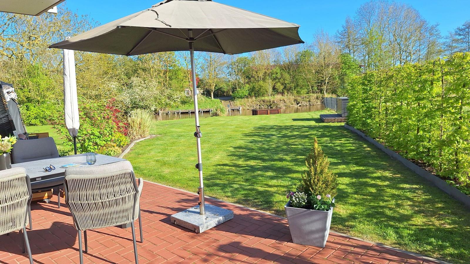 Terrasse mit Sonnenschirm, Tisch und Stühlen. Grasfläche, Bäume und Wasser im Hintergrund.