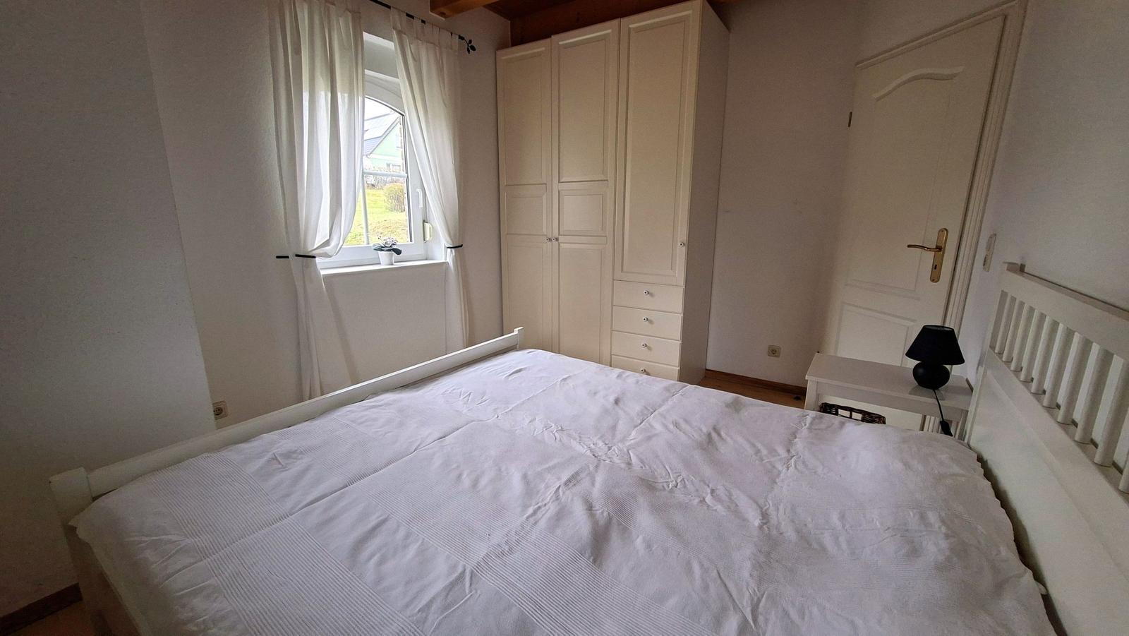 Bedroom with white bed, wardrobe, and window with white curtains.