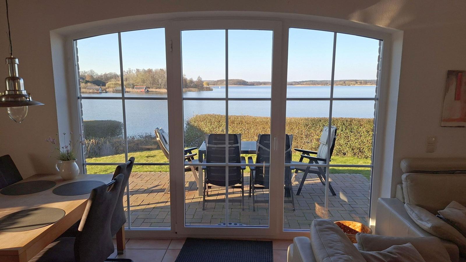Large window view of a lake. Terrace with table and chairs. Indoor area with dining table and sofa.