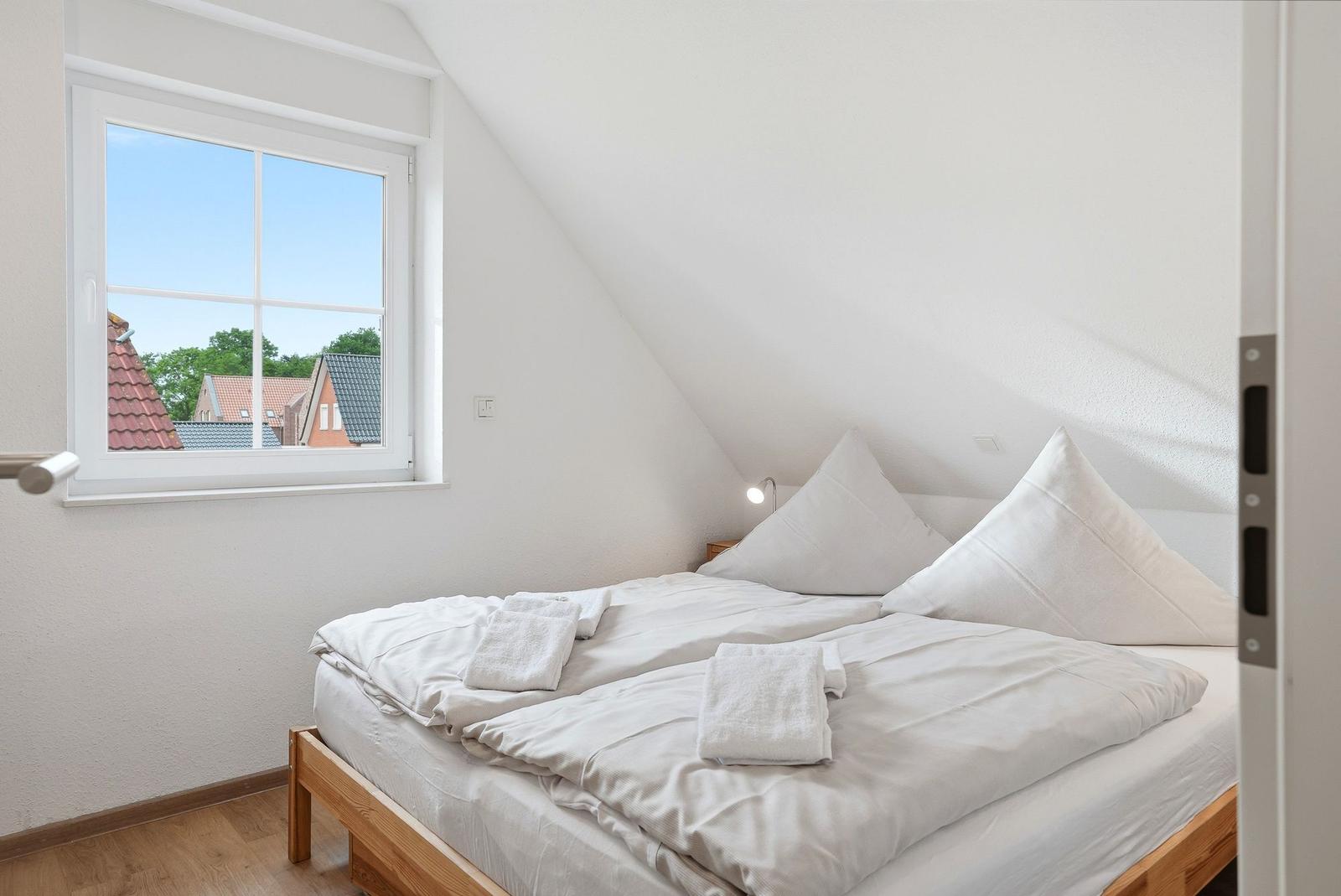 Schlafzimmer mit Dachschrägen, weißem Bett und Fensterblick auf Dächer.