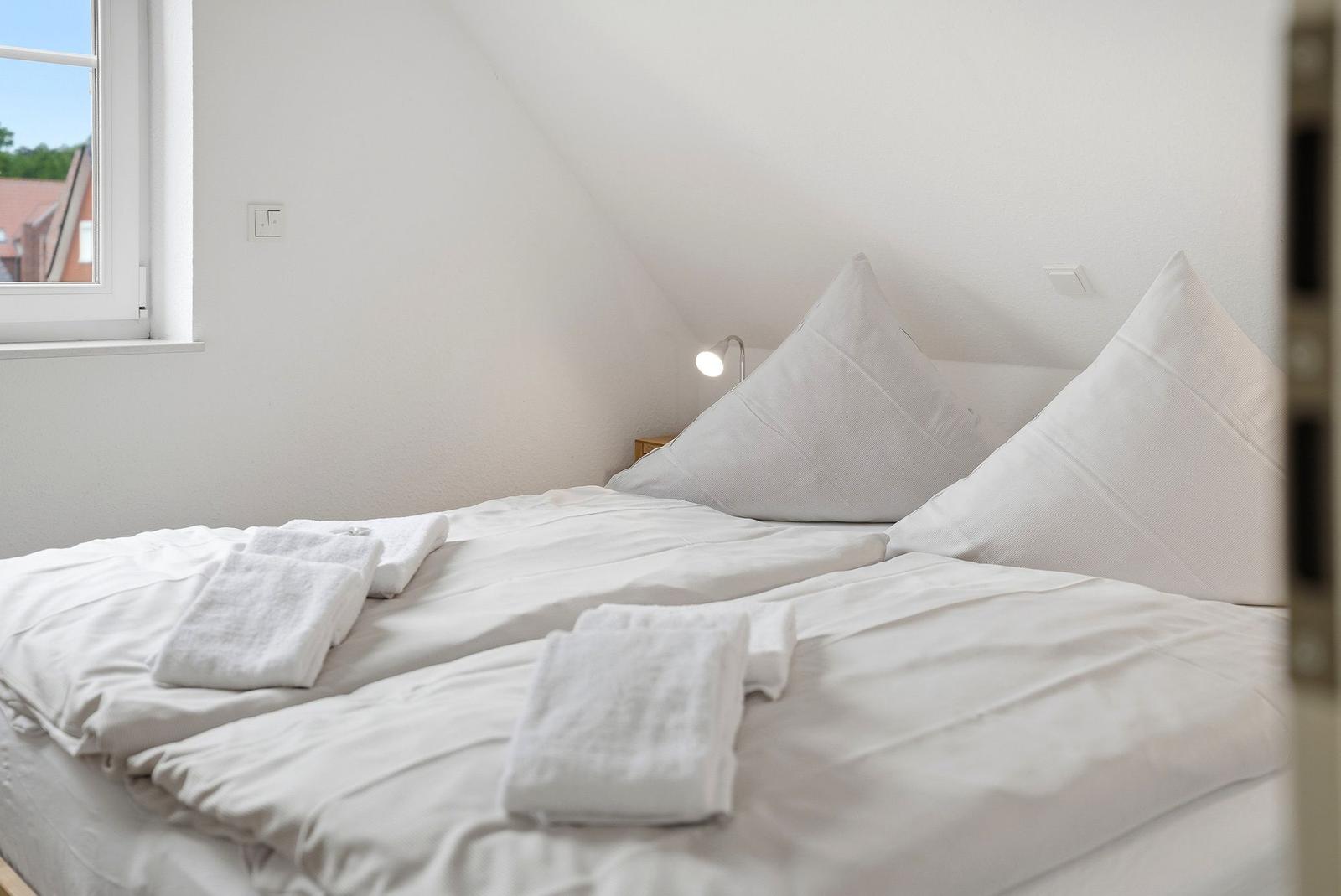 Ein Bett mit weißen Laken und Kissen. Auf dem Bett liegen gefaltete Handtücher. An der Wand hängt eine Lampe.