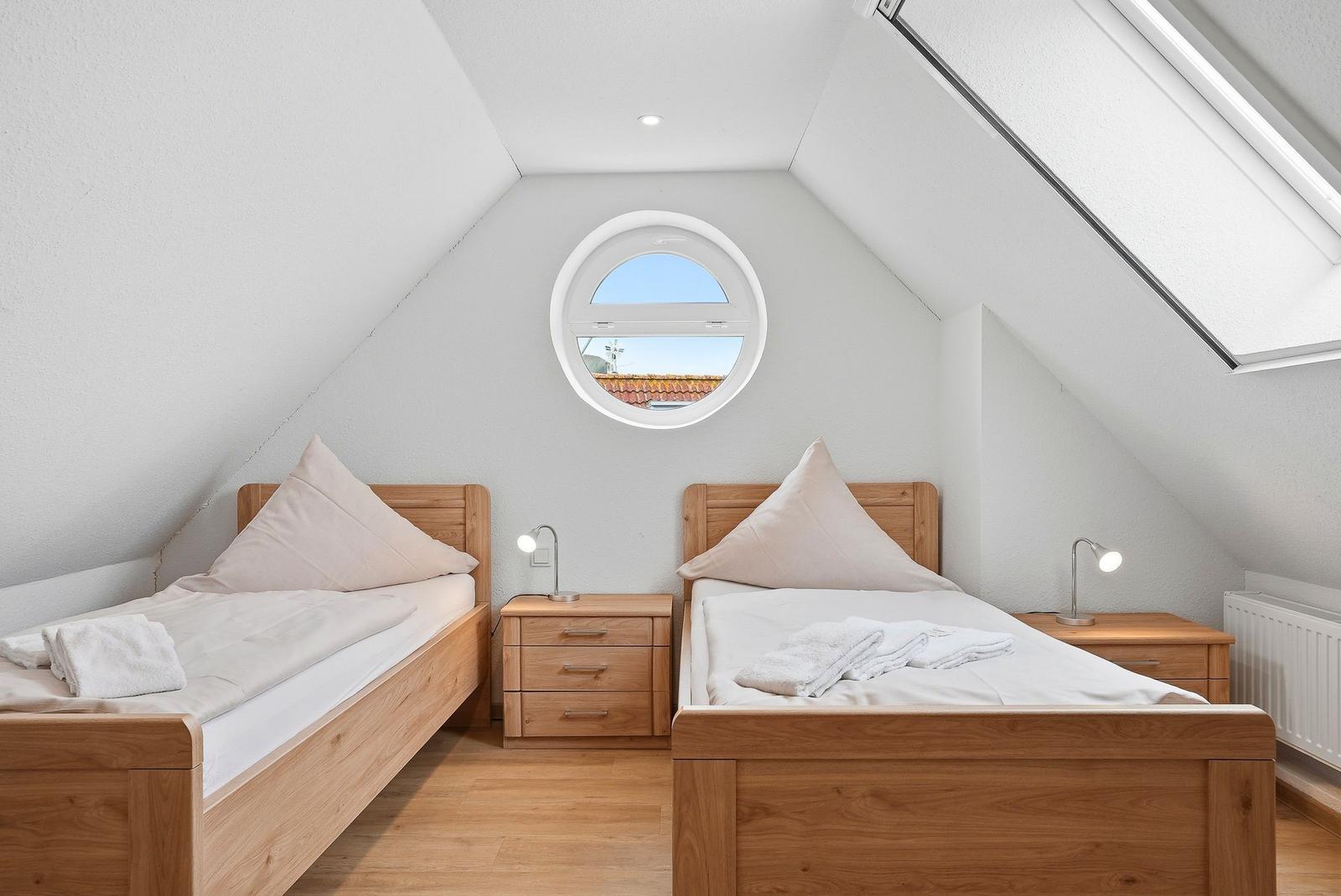 Zwei Einzelbetten mit Holzgestell, Nachttische und runde Fenster in Dachgeschosszimmer.