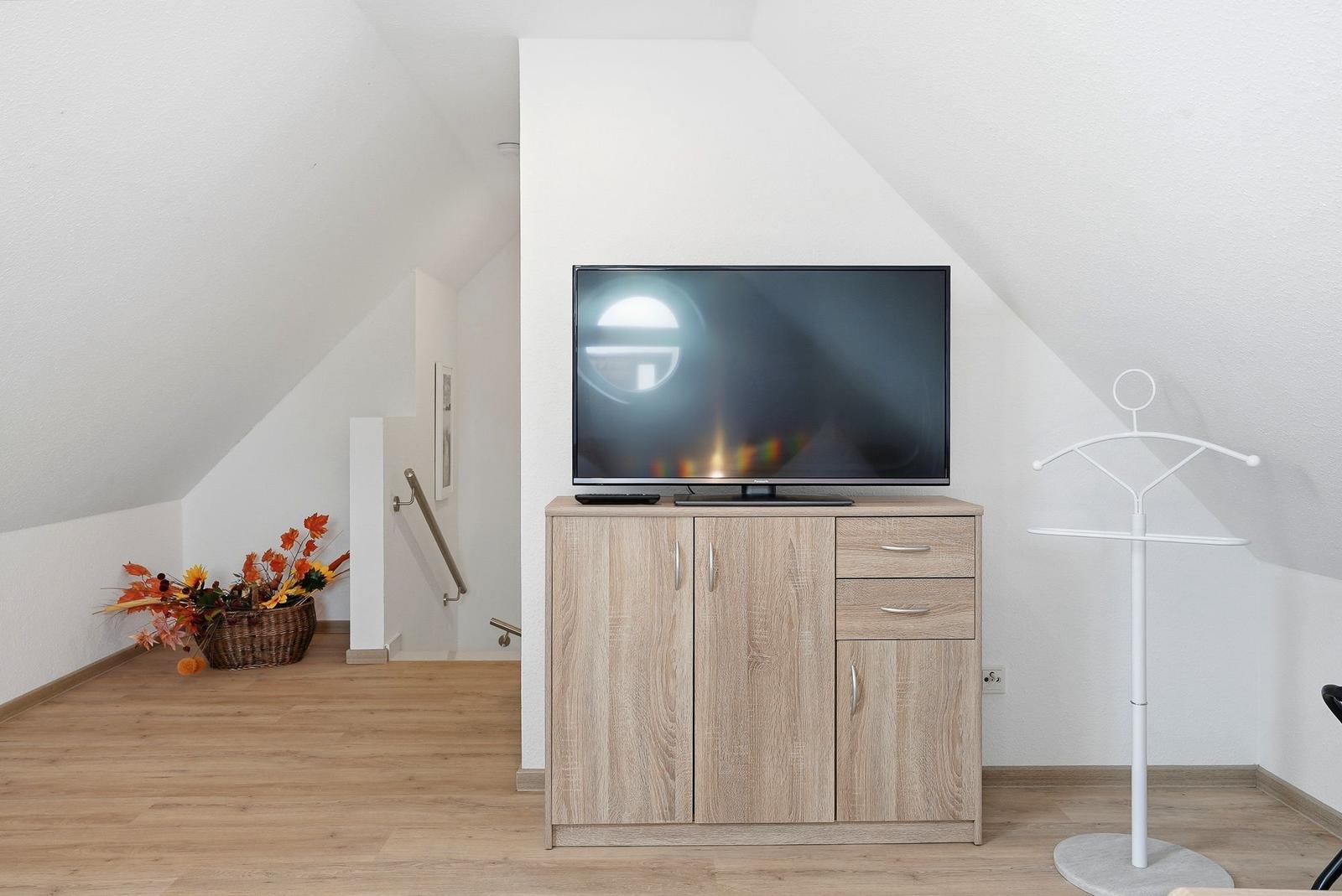 TV auf Holzschrank in Dachgeschosszimmer mit Kleiderständer und Korb mit Herbstblumen.