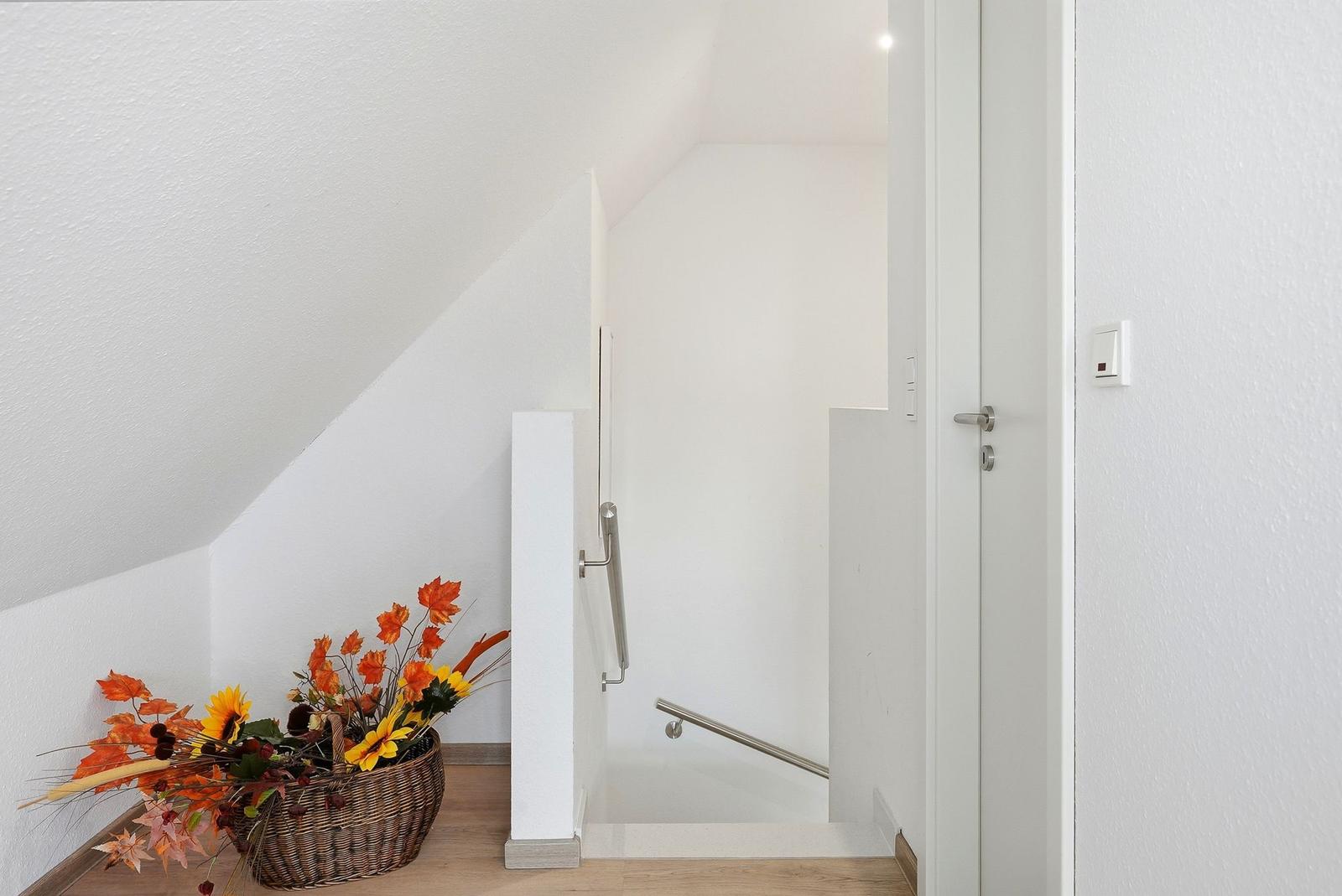 Hinter einer Tür führt eine Treppe mit Handlauf nach oben. Links steht ein Korb mit Herbstblumen.