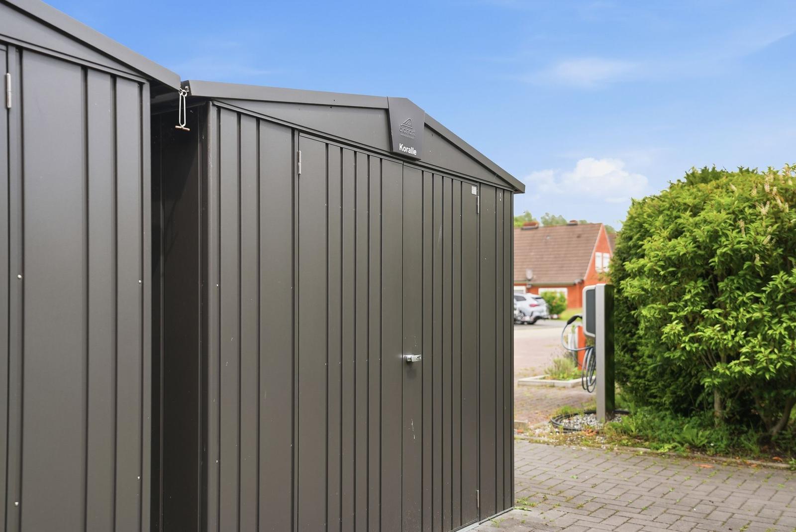 Graue Metallgarage mit Schriftzug Koralle. Hintergrund: Haus, Straße, grüne Hecke.