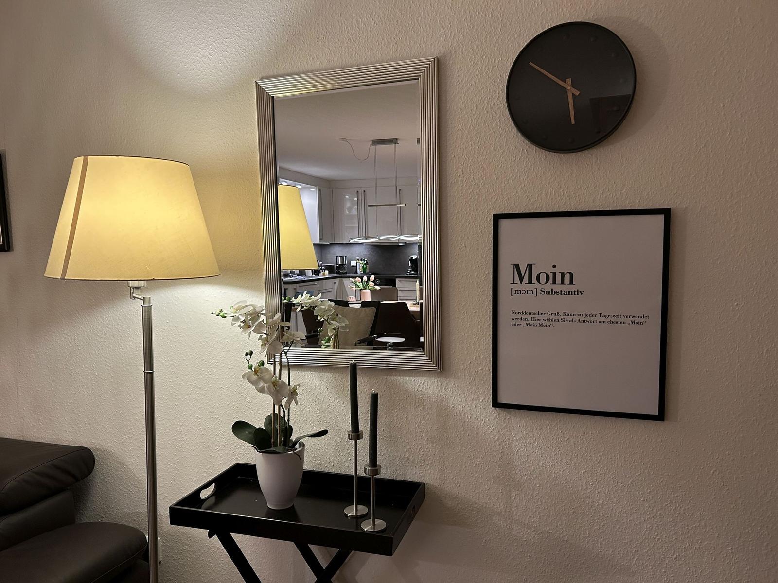 Wand mit Spiegel, Uhr und Bild. Tisch mit Pflanze und Kerzenhaltern.