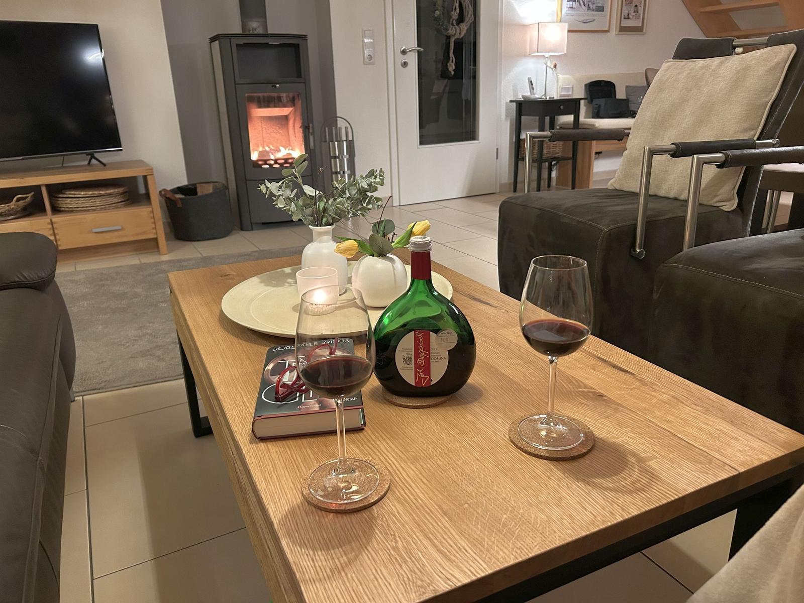 Holztisch mit Wein, Buch und Blumen. Kamin und Sofa im Hintergrund.