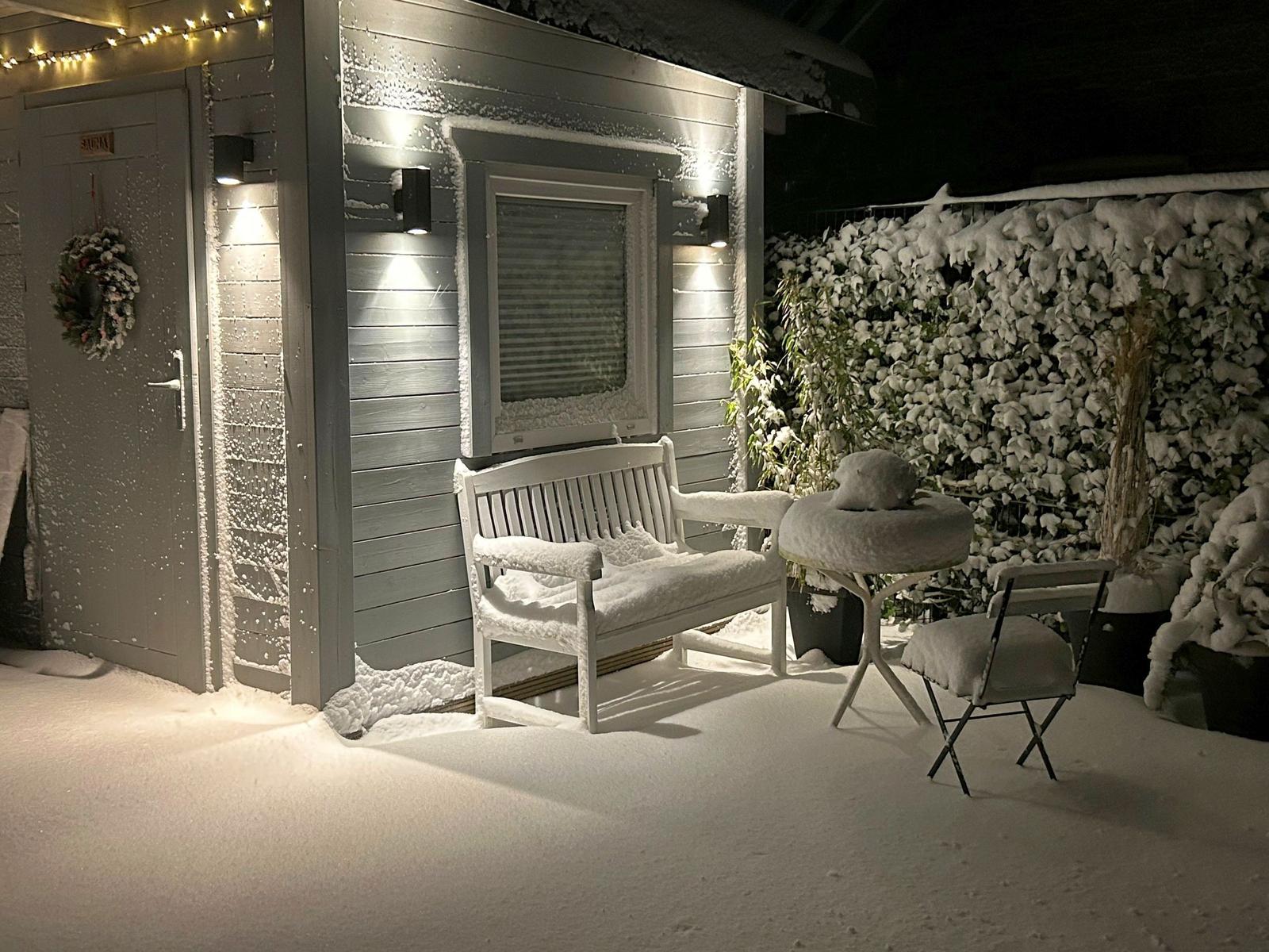 Schneebedeckter Außenbereich mit Bank, Tisch und Stuhl vor einem Haus mit Lichtern.