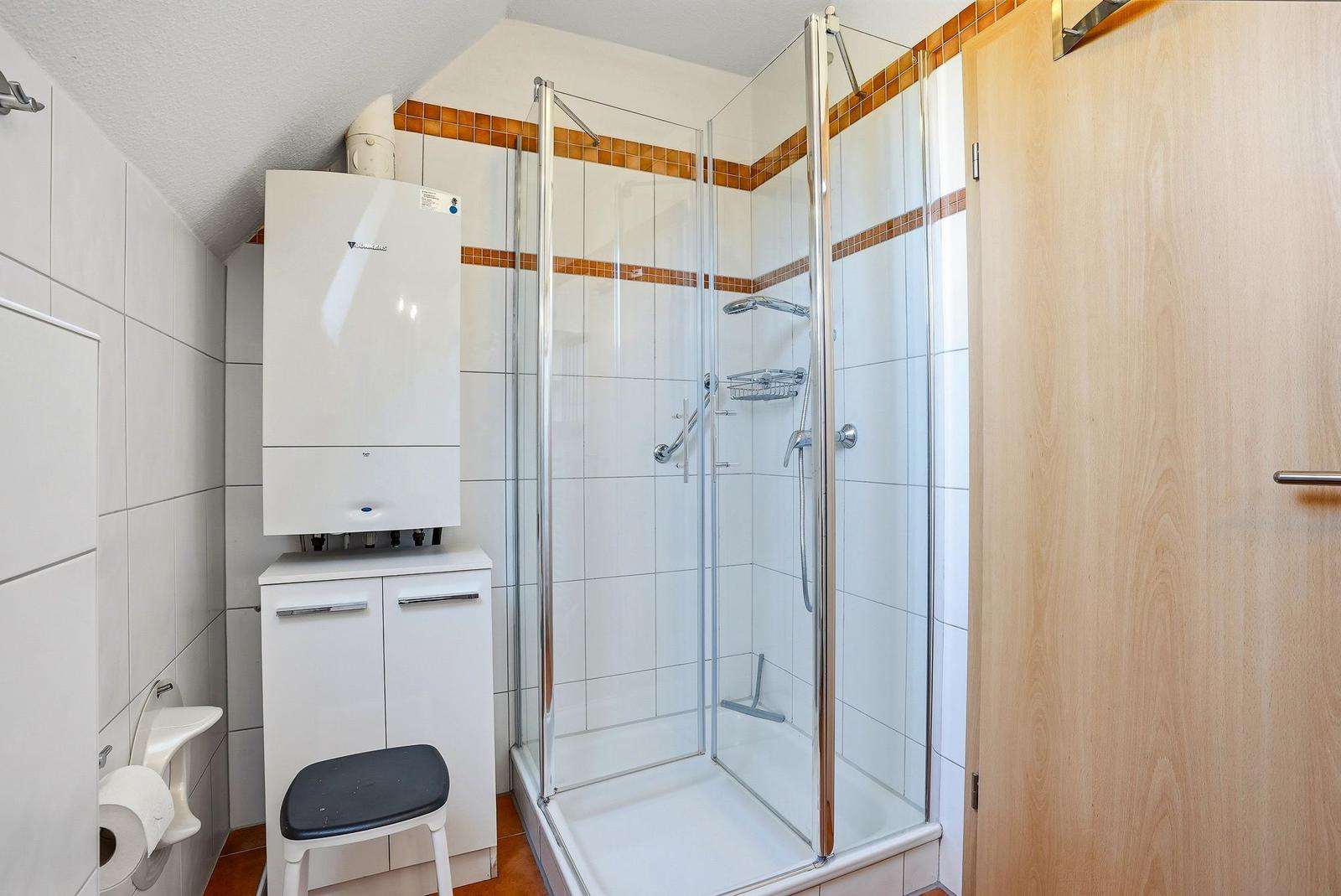 Badezimmer mit Dusche, Toilette und Heizung. Weiße Fliesen und Holztür.