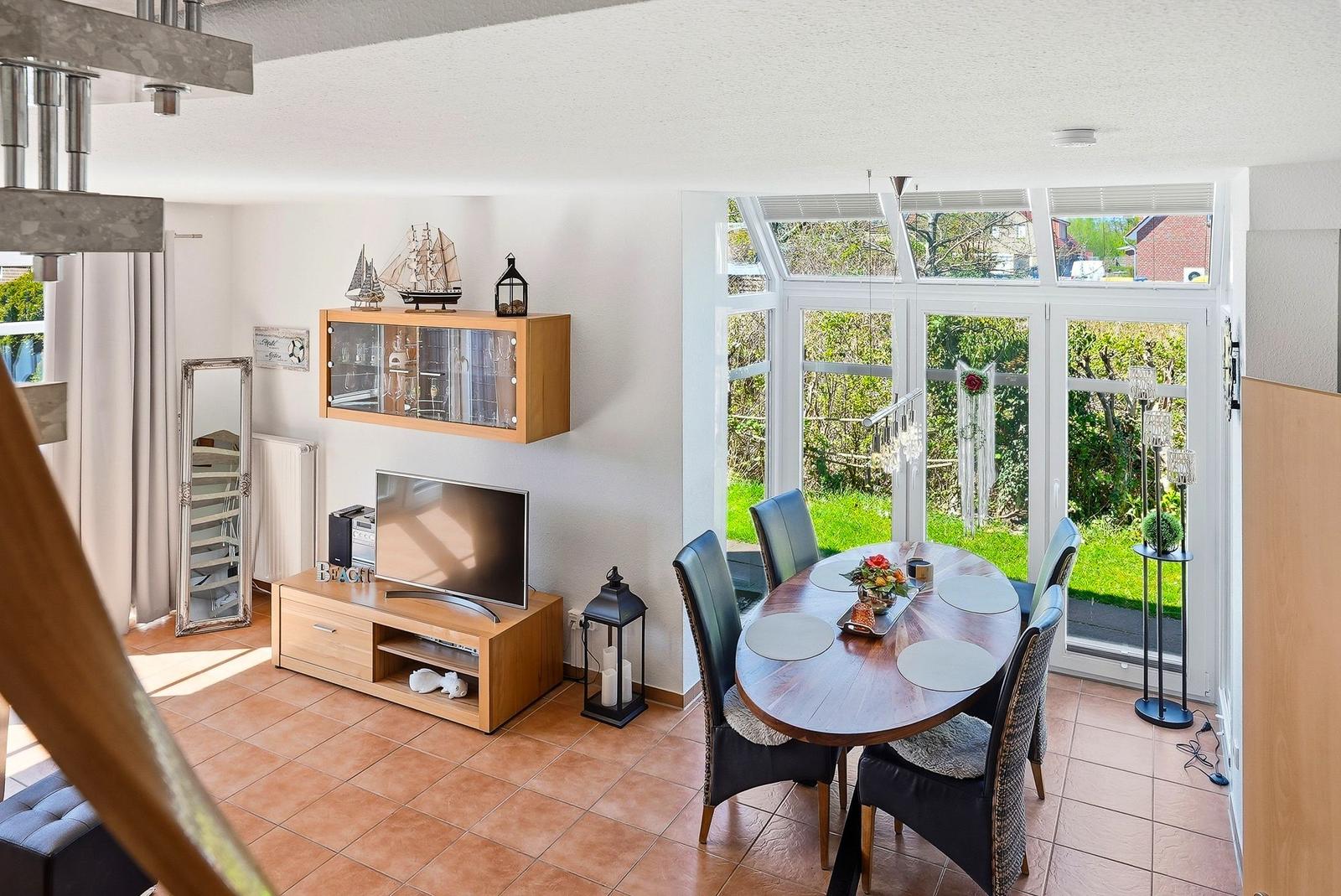 Esszimmer mit runden Tisch und Stühlen, Blick auf Garten. Wohnbereich mit Fernseher und Schrank.
