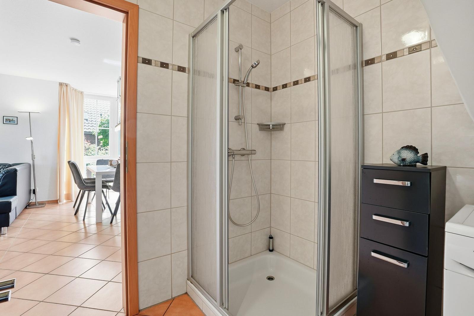 Dusche mit Duschbrause und Regal. Neben dem Bad ist ein Wohnzimmer mit Tisch und Stühlen.