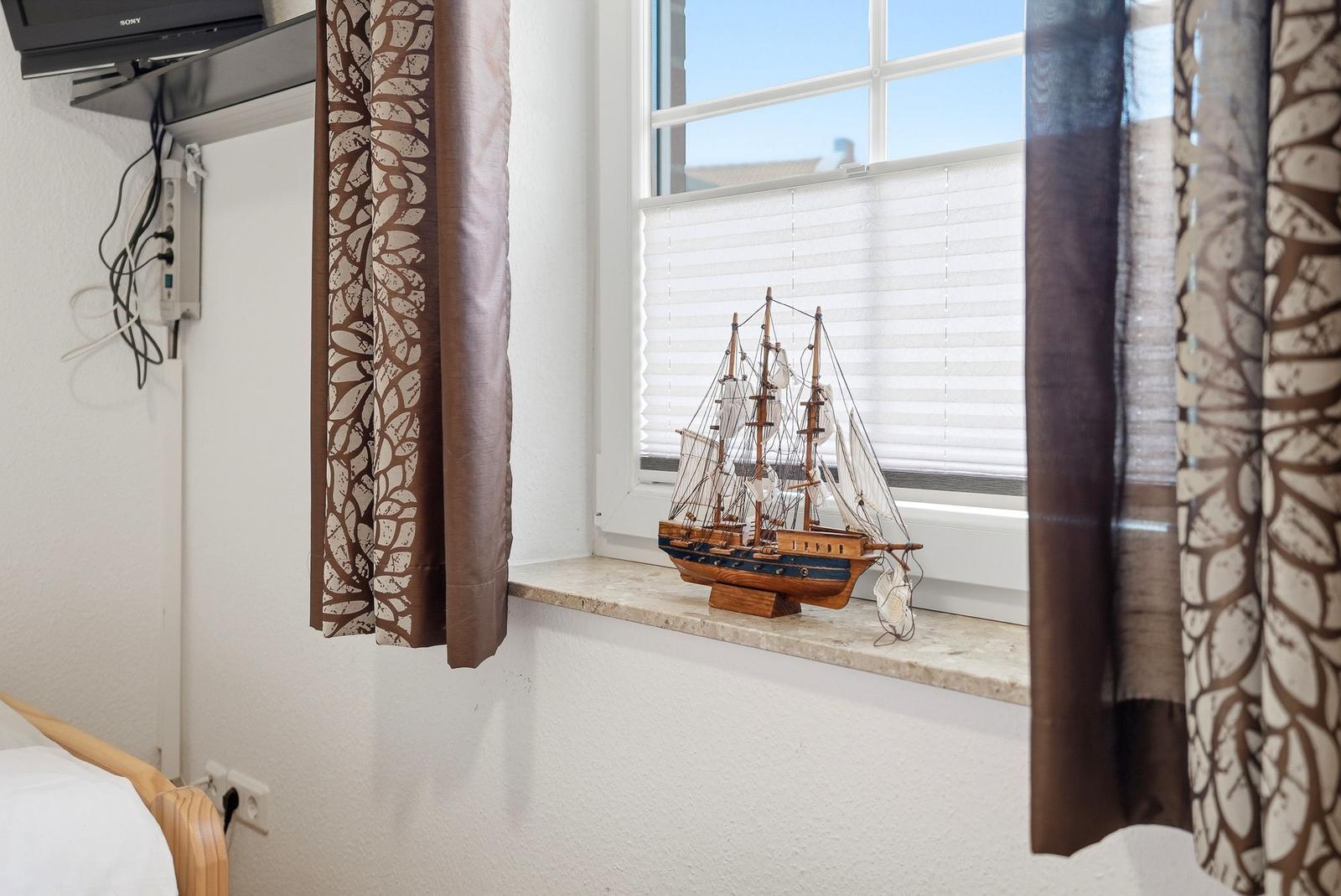 Ein Fenster mit Jalousie und Vorhang, auf dem Sims steht ein hölzernes Segelschiffmodell.