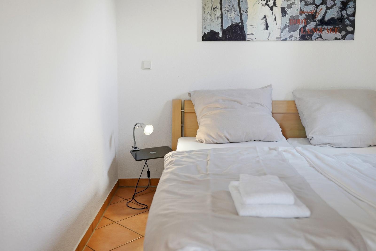Ein Bett mit weißen Laken und Handtüchern. An der Wand hängt ein Bild. Eine Lampe steht auf einem Nachttisch.