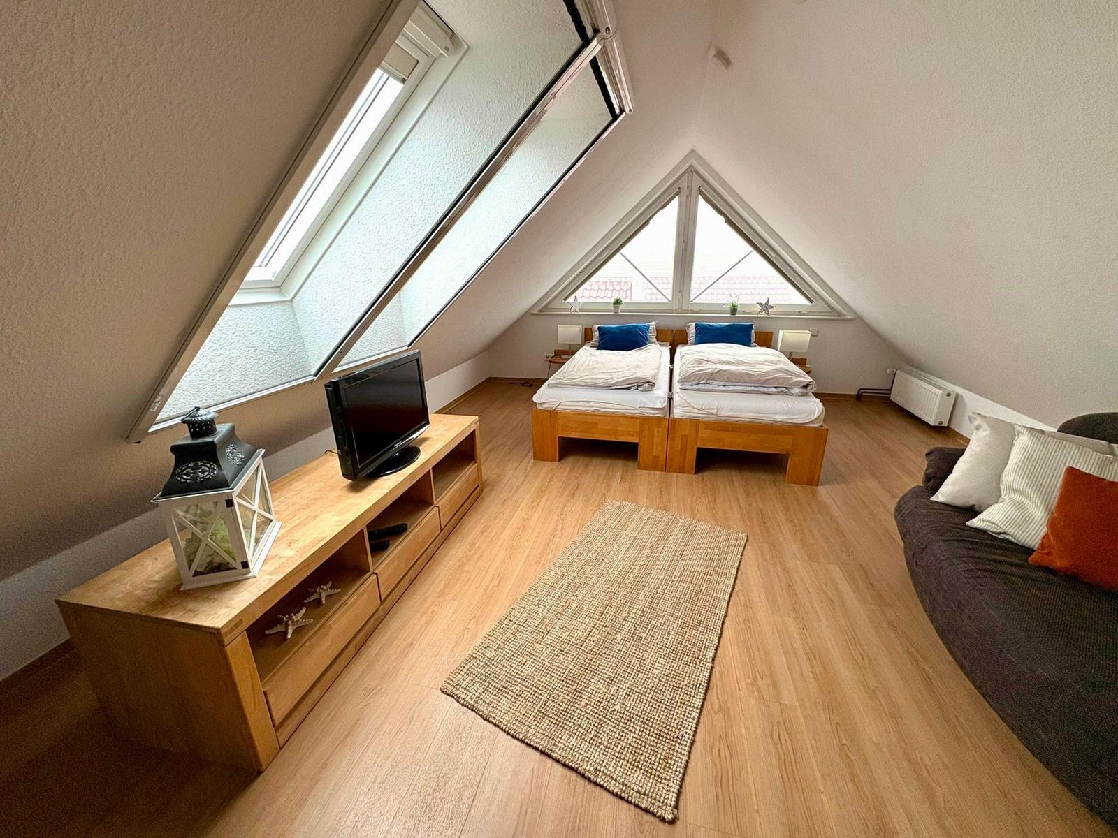 Zimmer mit zwei Betten, TV und großem Fenster. Holzboden und Sofa sichtbar.