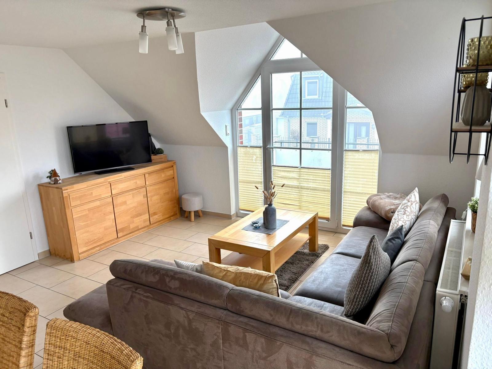 Wohnzimmer mit Sofa, Couchtisch und TV. Große Fensterfront mit Blick auf Balkon.