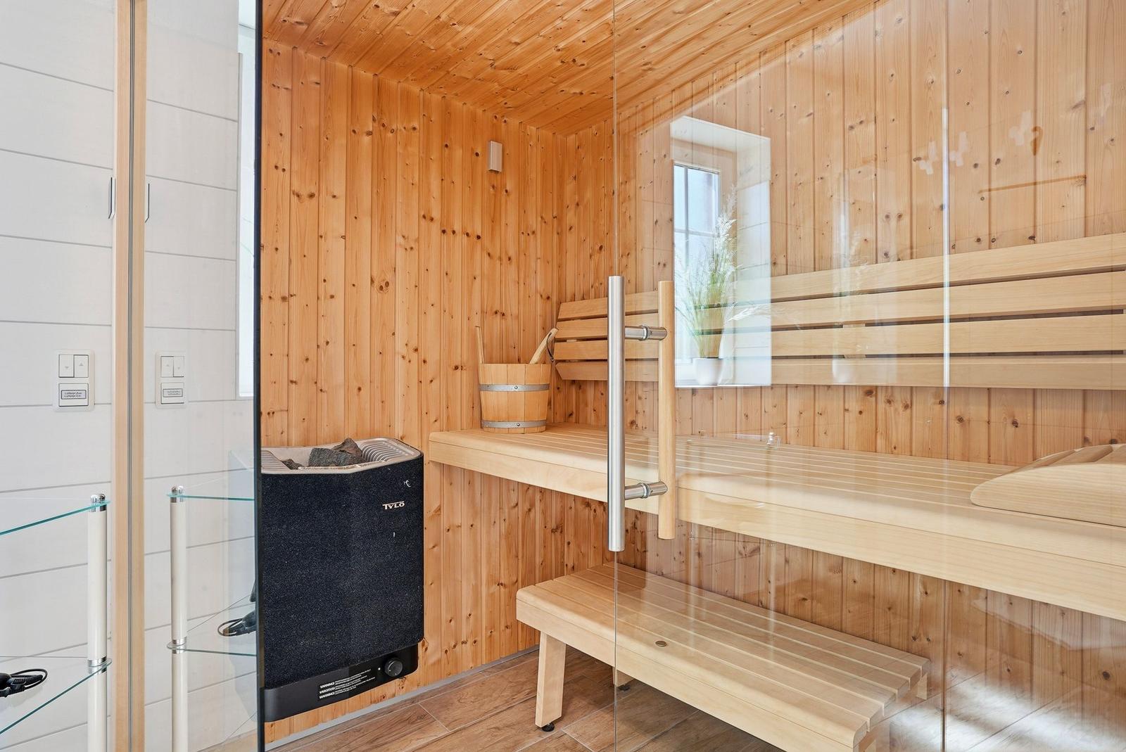 Sauna mit Holzwand, Bänken und Heizgerät. Glaswand und Fenster sichtbar.