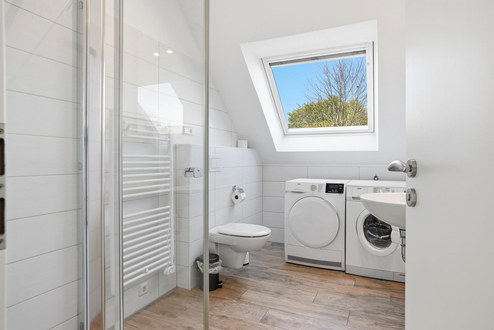 Badezimmer mit Dusche, Toilette, Waschmaschine und Wärmepumpe. Dachfenster mit Blick auf Bäume.
