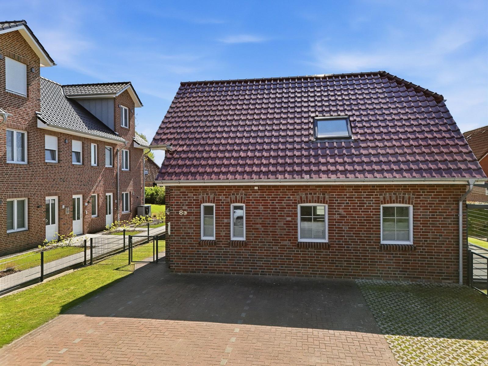 Ein zweistöckiges Haus mit rotem Ziegeldach und weißen Fenstern. Vor dem Haus befindet sich ein gepflasterter Hof.