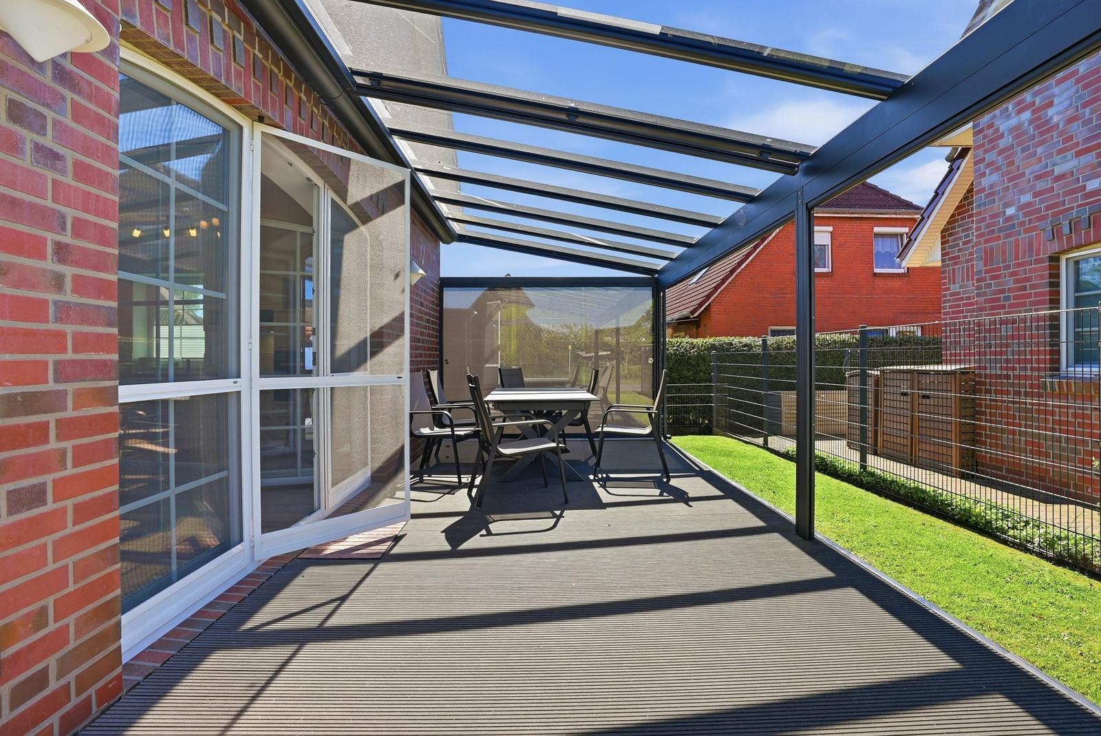 Gedeckter Balkon mit Tisch und Stühlen. Glasdach und Holzboden. Grünfläche im Hintergrund.