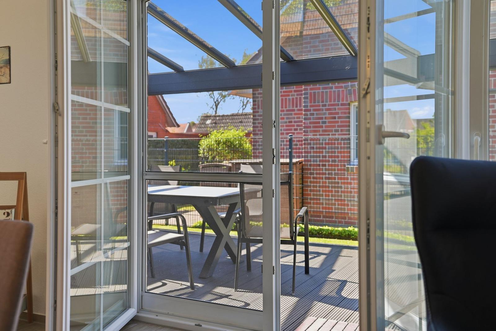 Durchgehende Glastüren führen auf eine Terrasse mit Tisch und Stühlen unter einer Dachkonstruktion.