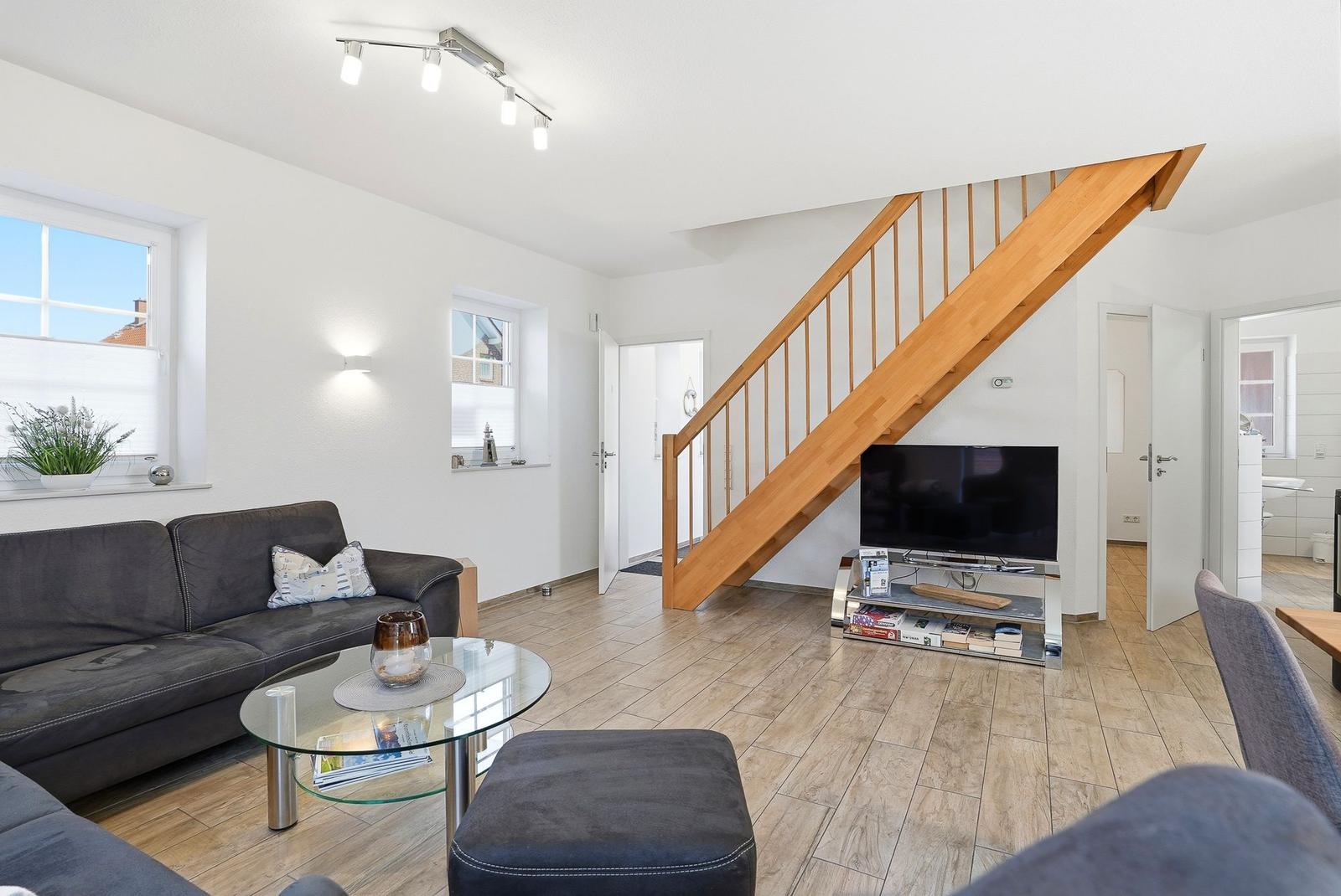 Wohnzimmer mit Sofa, Glas-Tisch und Holztreppe. Fernseher und Fenster sichtbar.