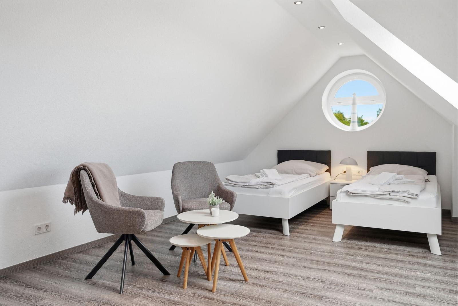 Two beds, two chairs, and a round window view in an attic room.