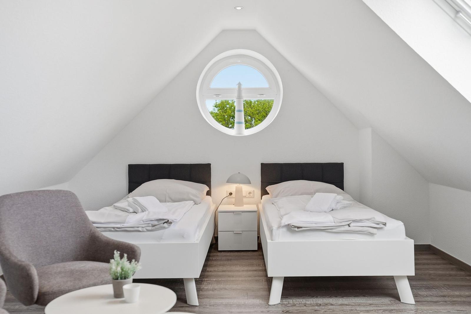 Two beds under sloped ceilings with round window. Wooden floor and gray seating area.