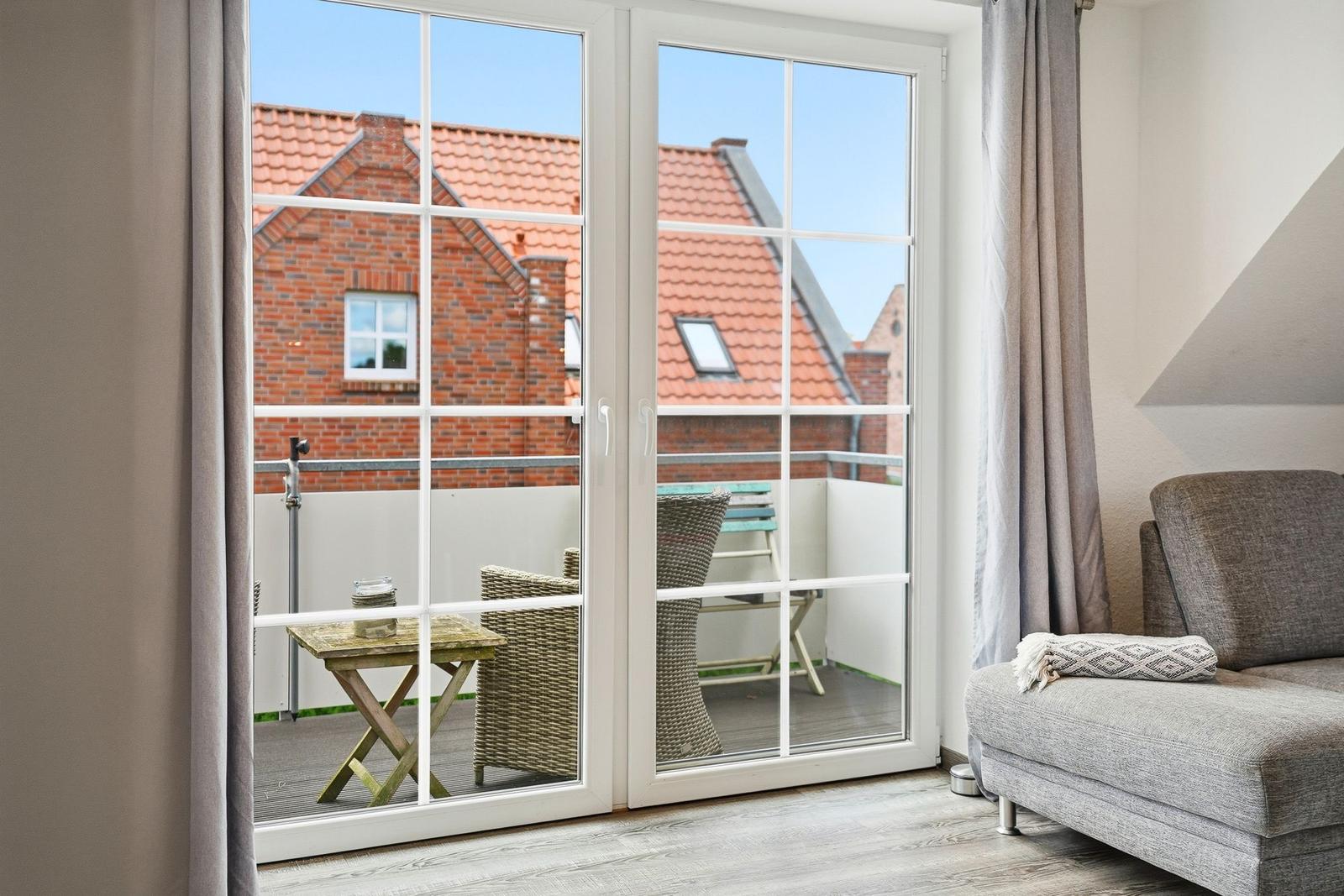 Through glass doors, a balcony with table and chairs is visible. Red roofs are in the background.