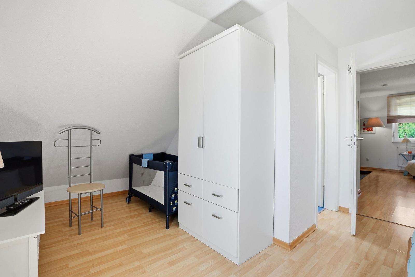 Schlafzimmer mit weißem Schrank, Kinderbett und Fernseher. Holzboden und angrenzender Flur.