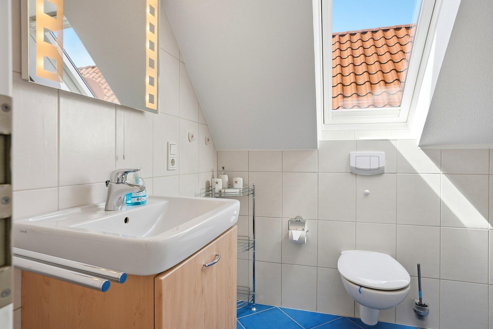 Badezimmer mit Waschbecken, Spiegel und Toilette. Dachfenster mit Blick auf Dachziegel.