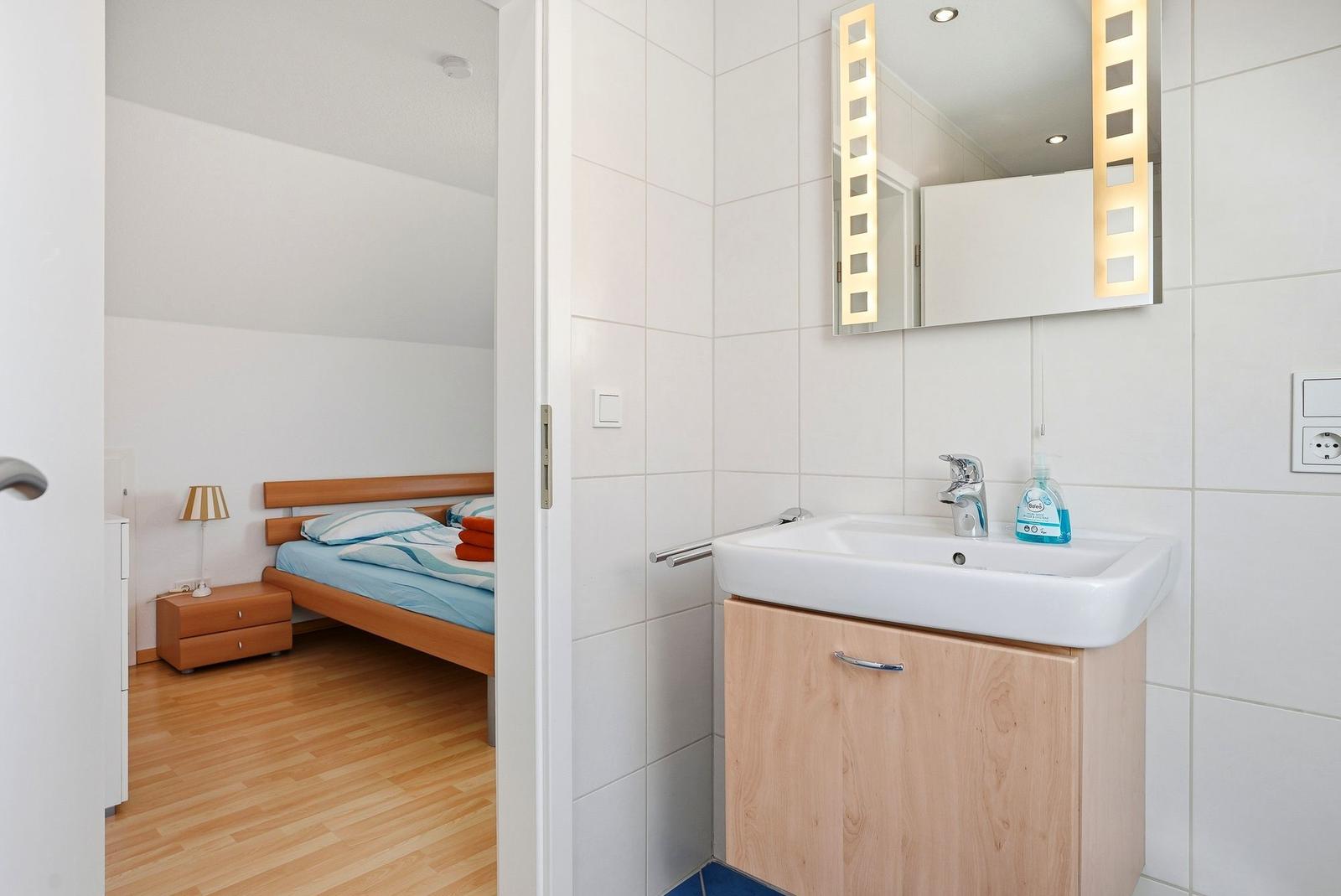 Badezimmer mit Waschbecken und Spiegel. Im Hintergrund ist ein Schlafzimmer mit Bett und Nachttisch sichtbar.