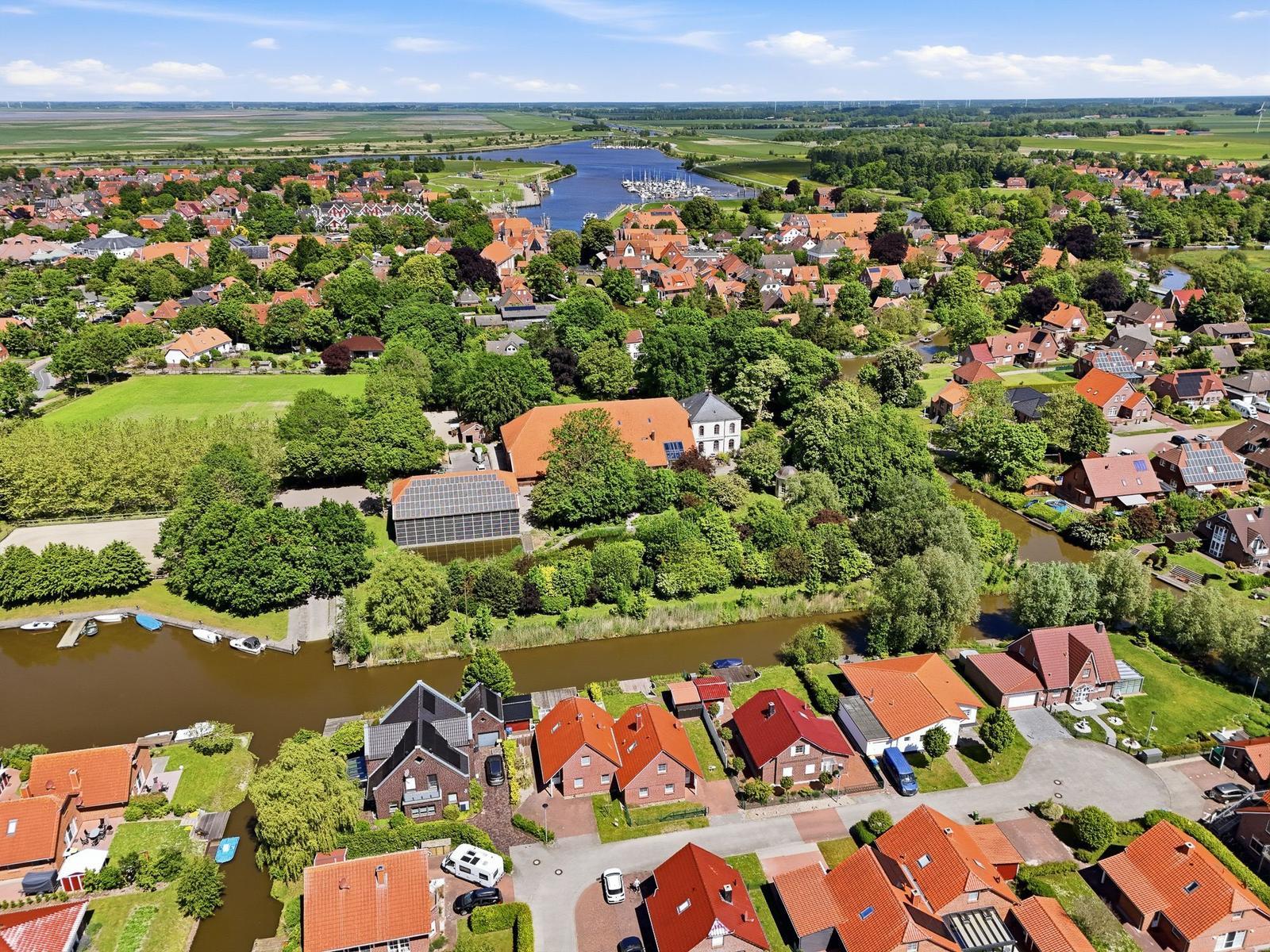 Luftbild von Häusern mit roten Dächern am Fluss. Grünflächen und Boote sind sichtbar.