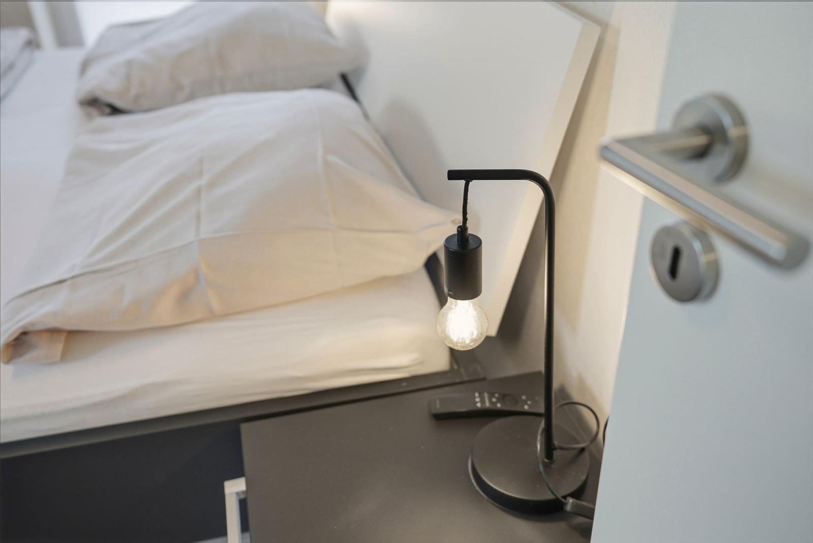 Schlafzimmer mit Bett, Nachttisch und Lampe. Tür mit Griff und Schlüsselloch sichtbar.