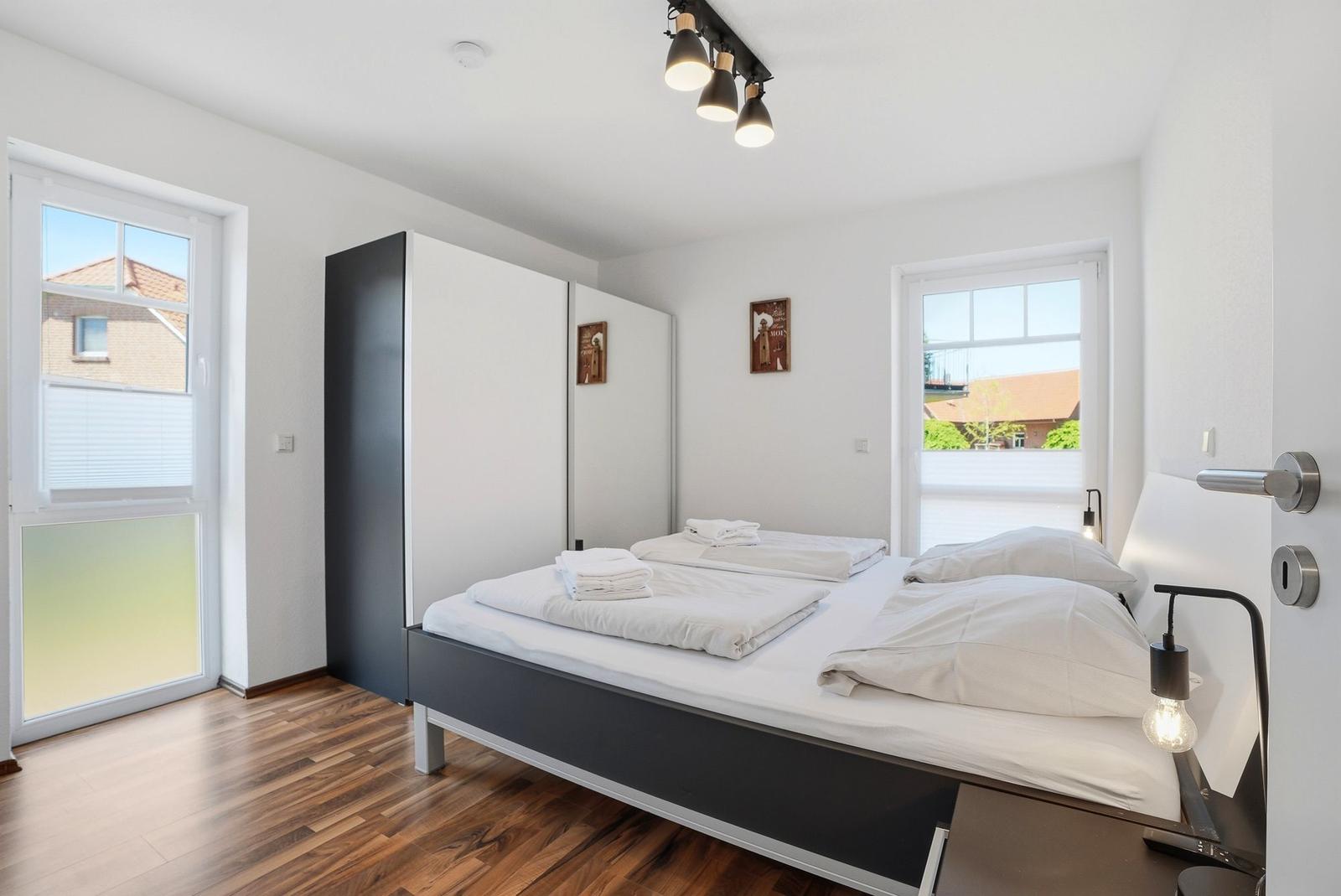 Schlafzimmer mit Doppelbett, zwei Fenstern und Holzboden. Neben dem Bett steht ein Schrank.