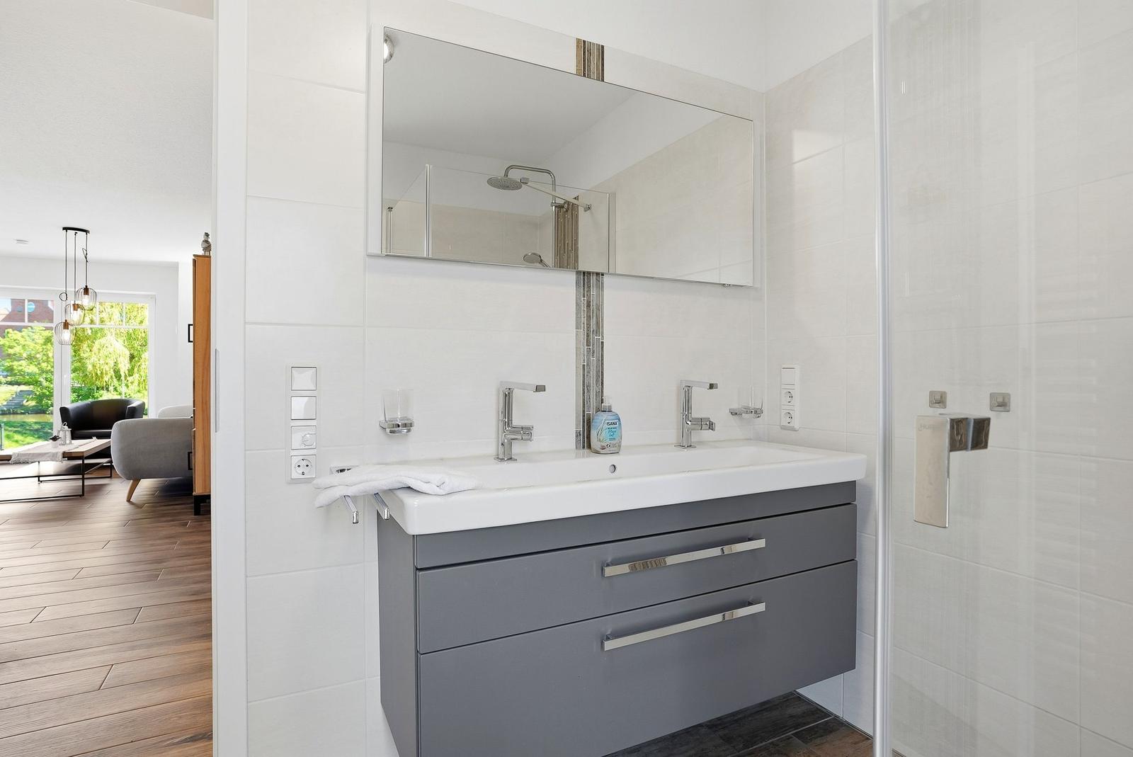 Badezimmer mit Waschtisch, Spiegel und Dusche. Blick in Wohnbereich mit Sofa.