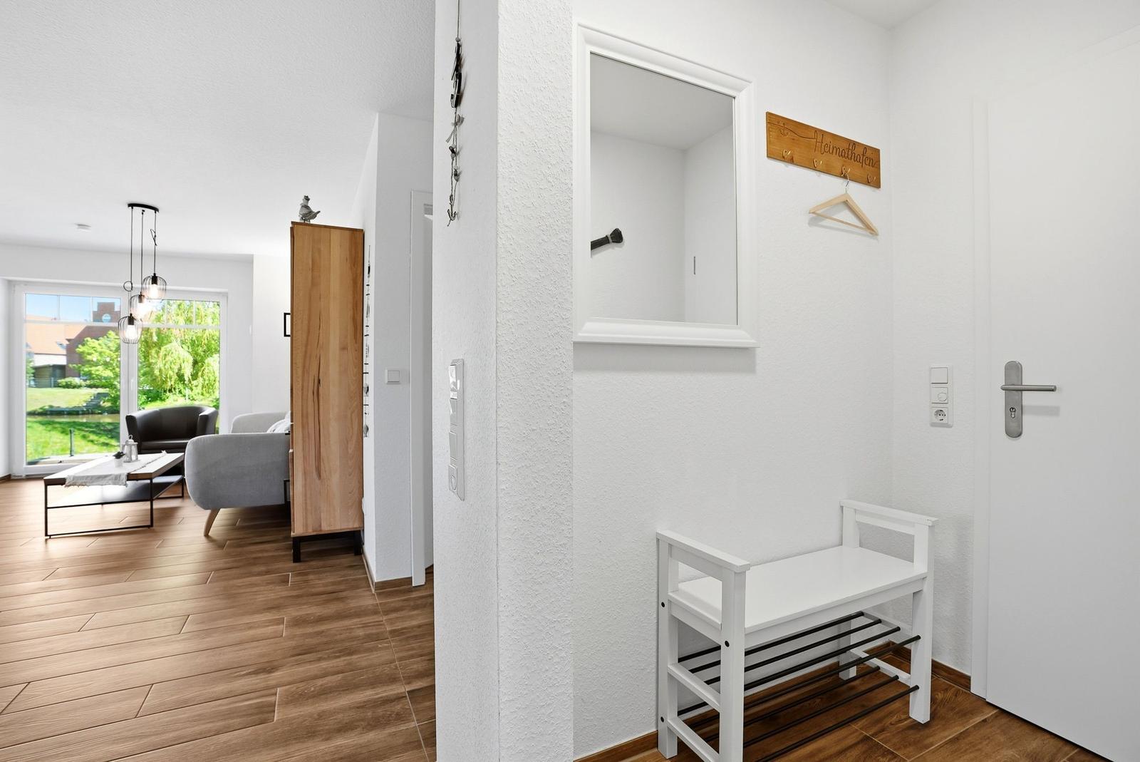 Eingangsbereich mit Spiegelschrank, Hocker und Holzfußboden. Blick ins Wohnzimmer mit Sofa und Fenster.