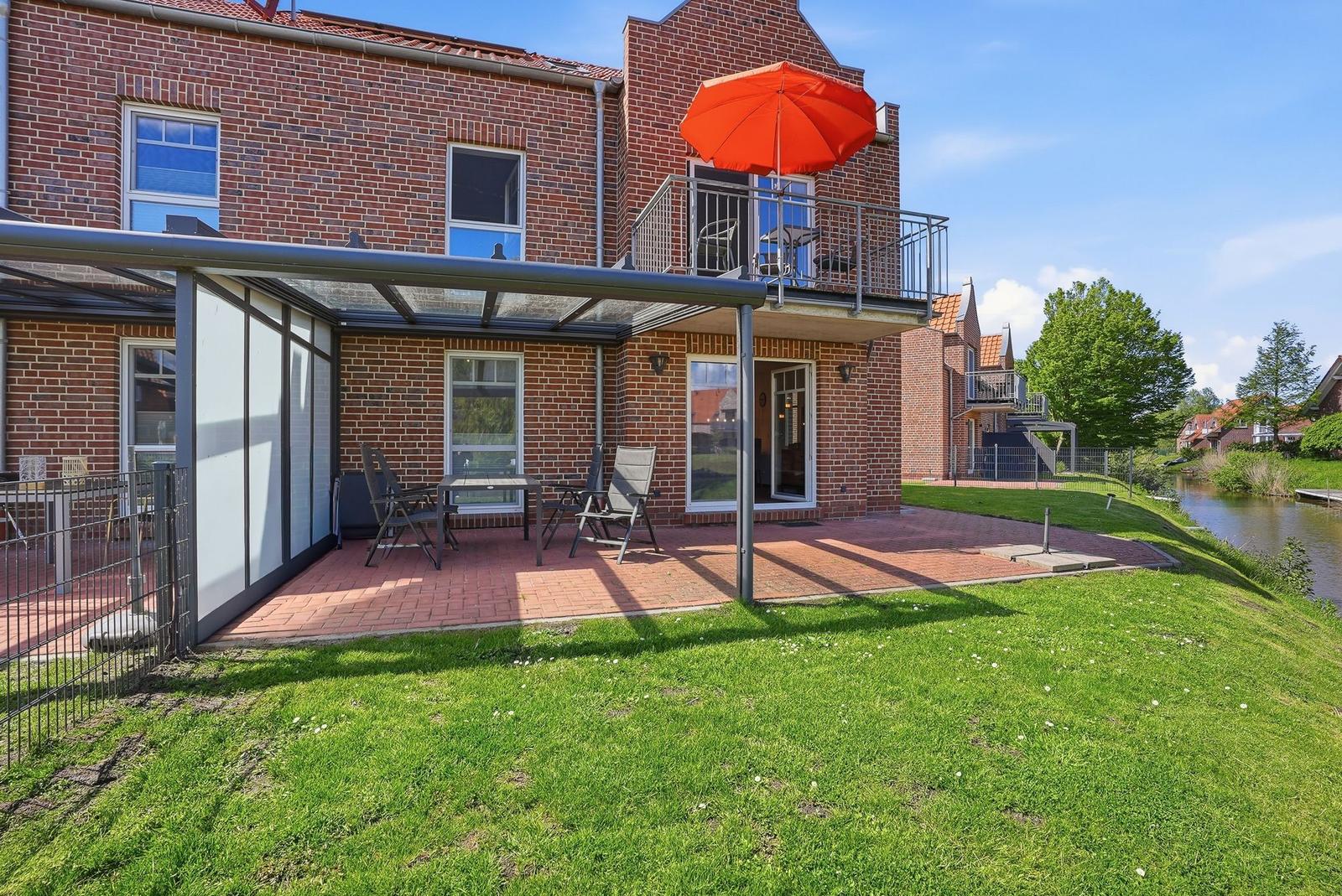 Rotes Ziegelhaus mit Terrasse und orangefarbener Sonnenschirm. Grünfläche und Wasser im Hintergrund.
