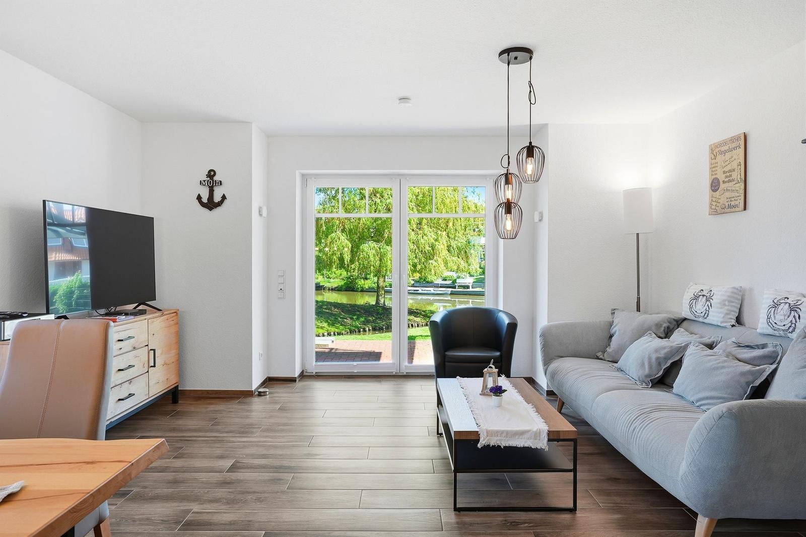 Wohnzimmer mit Sofa, Sessel und Fernseher. Durchgang zur Terrasse mit Blick auf Wasser.