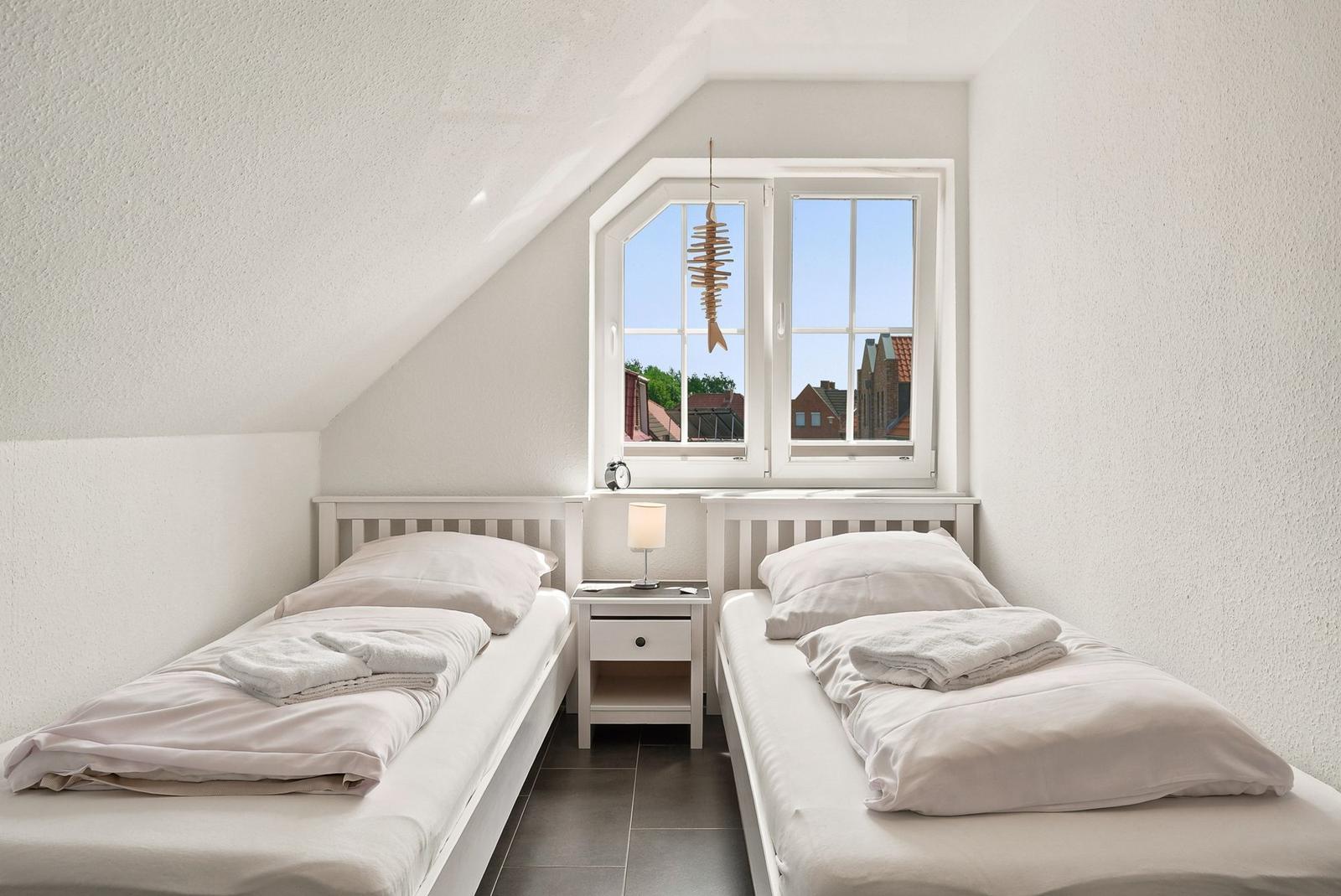 Zwei Einzelbetten mit weißer Bettwäsche stehen unter einem Dachfenster mit Blick auf Häuser.