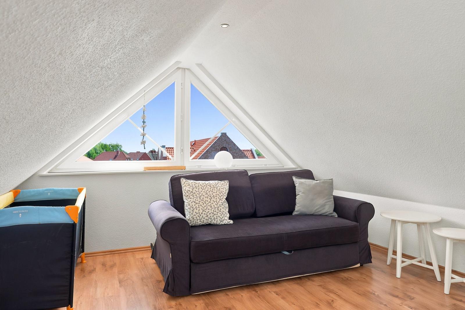 Dachgeschosswohnung mit Sofa, Babybett und Dachfenster. Blick auf Dächer und Grün.