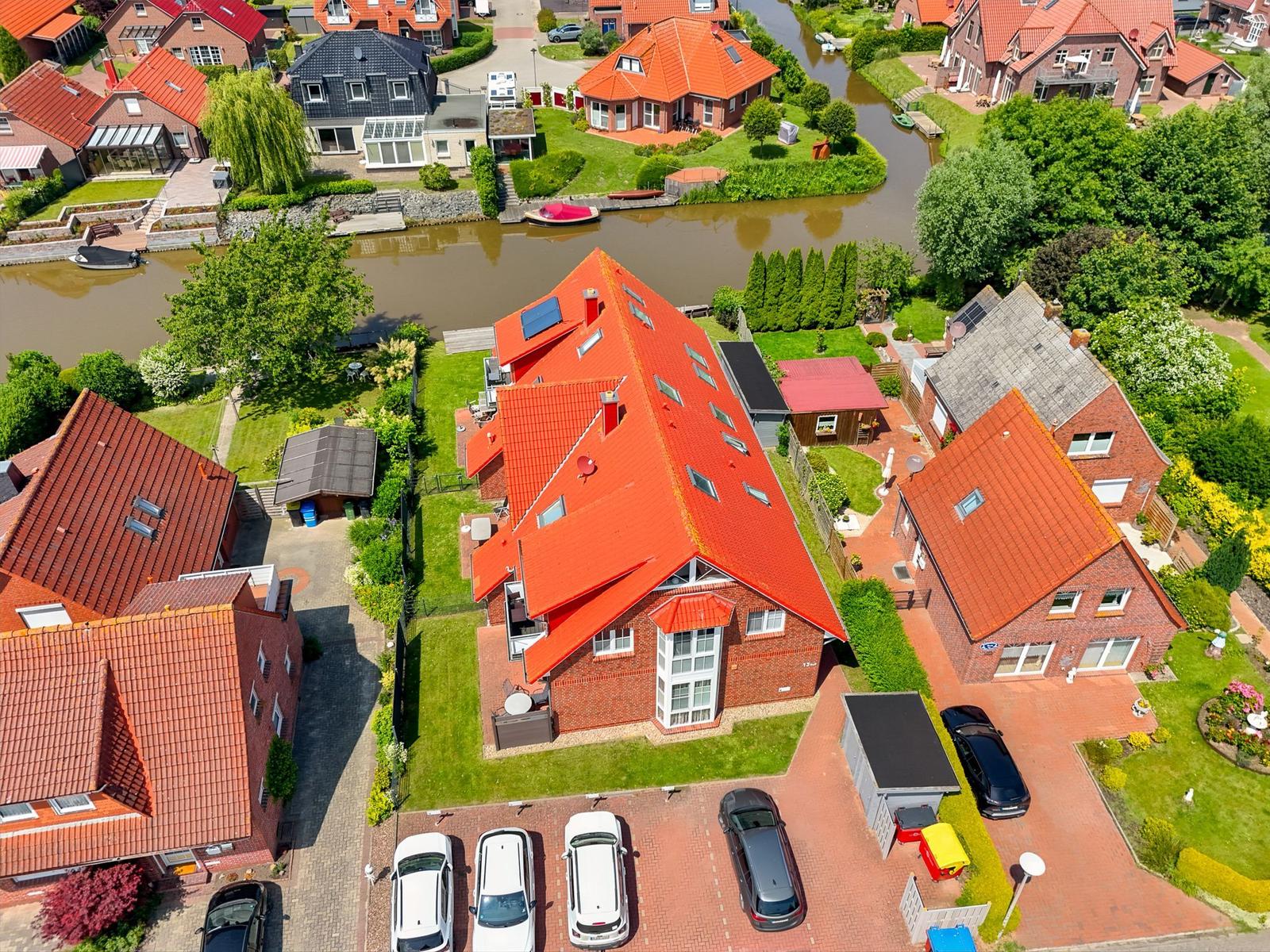 Haus mit rotem Dach und Garten an Fluss, mehrere Autos auf Hof.