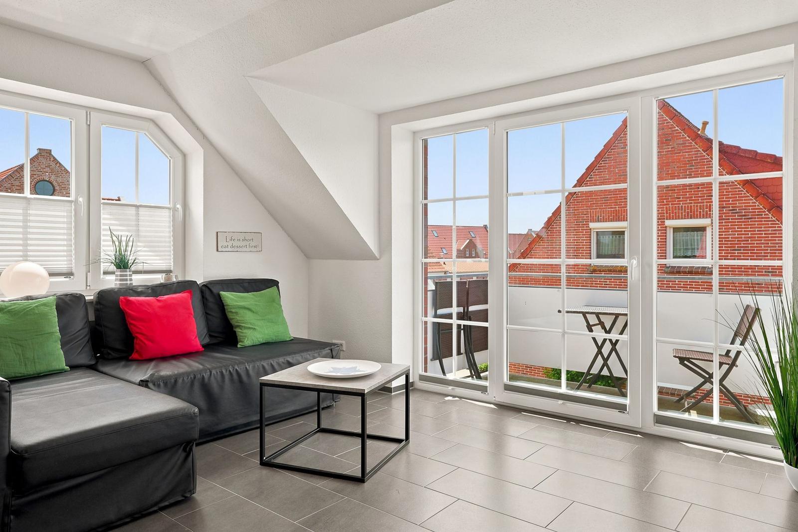 Wohnzimmer mit Sofa, Tisch und Balkon. Durchgehende Glasfront mit Blick auf Dächer.